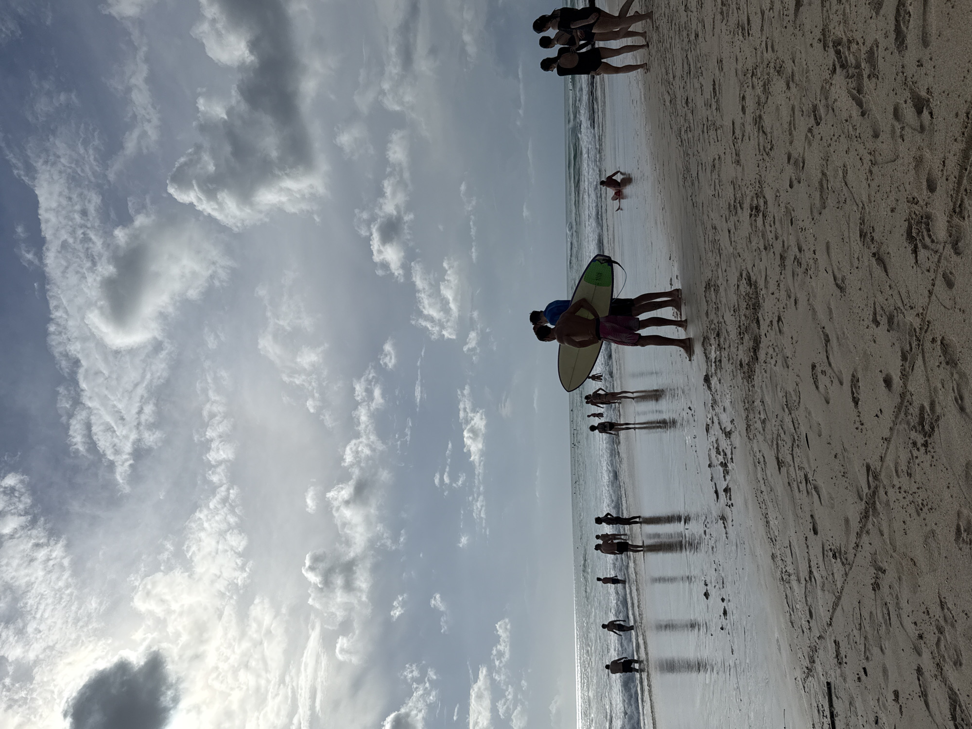The image depicts a beach in Kuta Selatan, Indonesia. The sky is filled with clouds. A group of people is walking along the shoreline, some in the water. Two individuals are carrying a surfboard. Footprints are visible in the sand.
