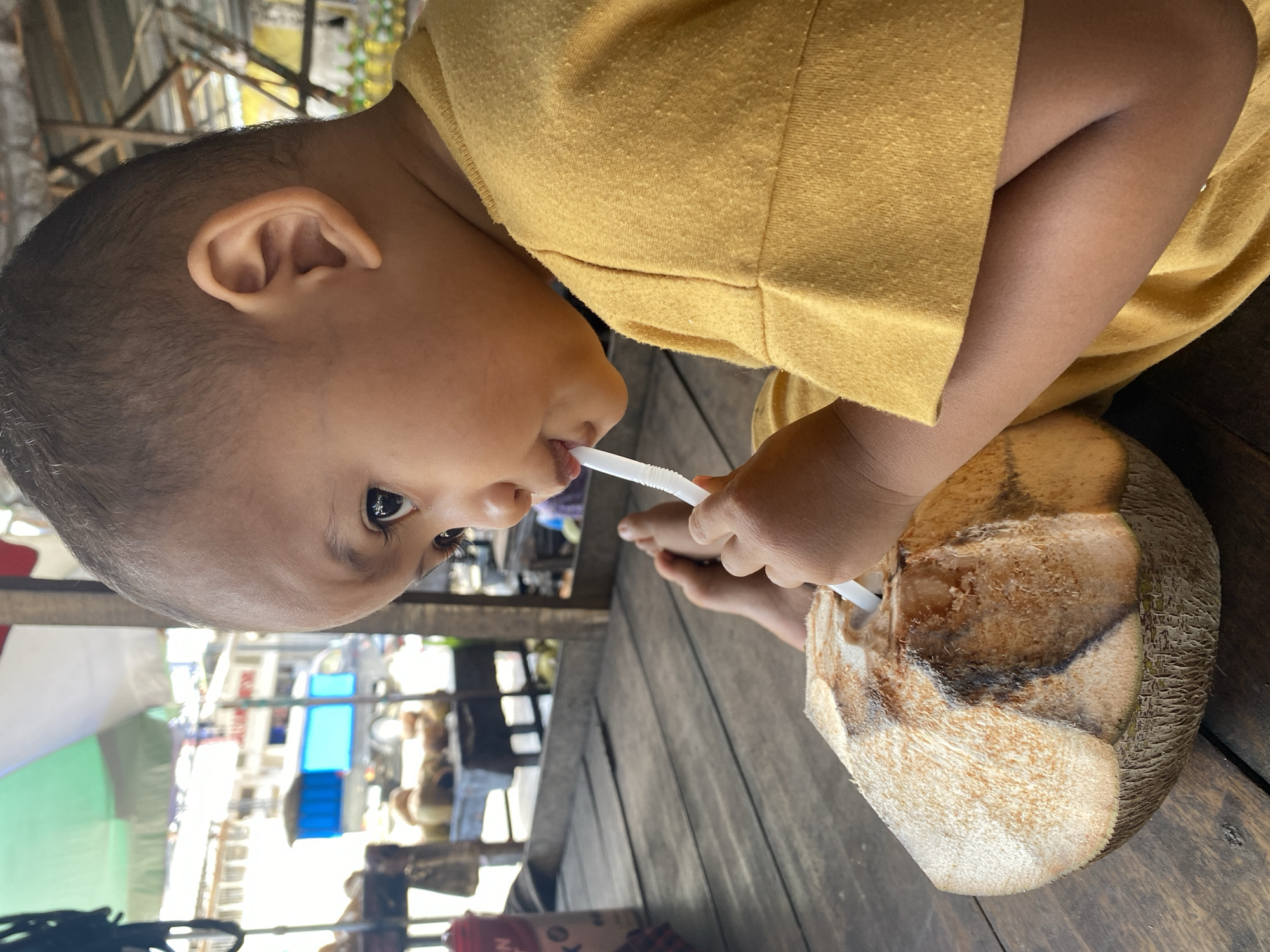 A young child, likely of Indonesian descent, is sitting on a wooden platform and drinking from a young coconut with a straw. The child is wearing a yellow t-shirt. The child's bare feet are visible. In the background, there are coconuts, market stalls, and a blue and white truck. The scene appears to be an outdoor market or roadside stall in Ta', Indonesia, likely during the daytime.