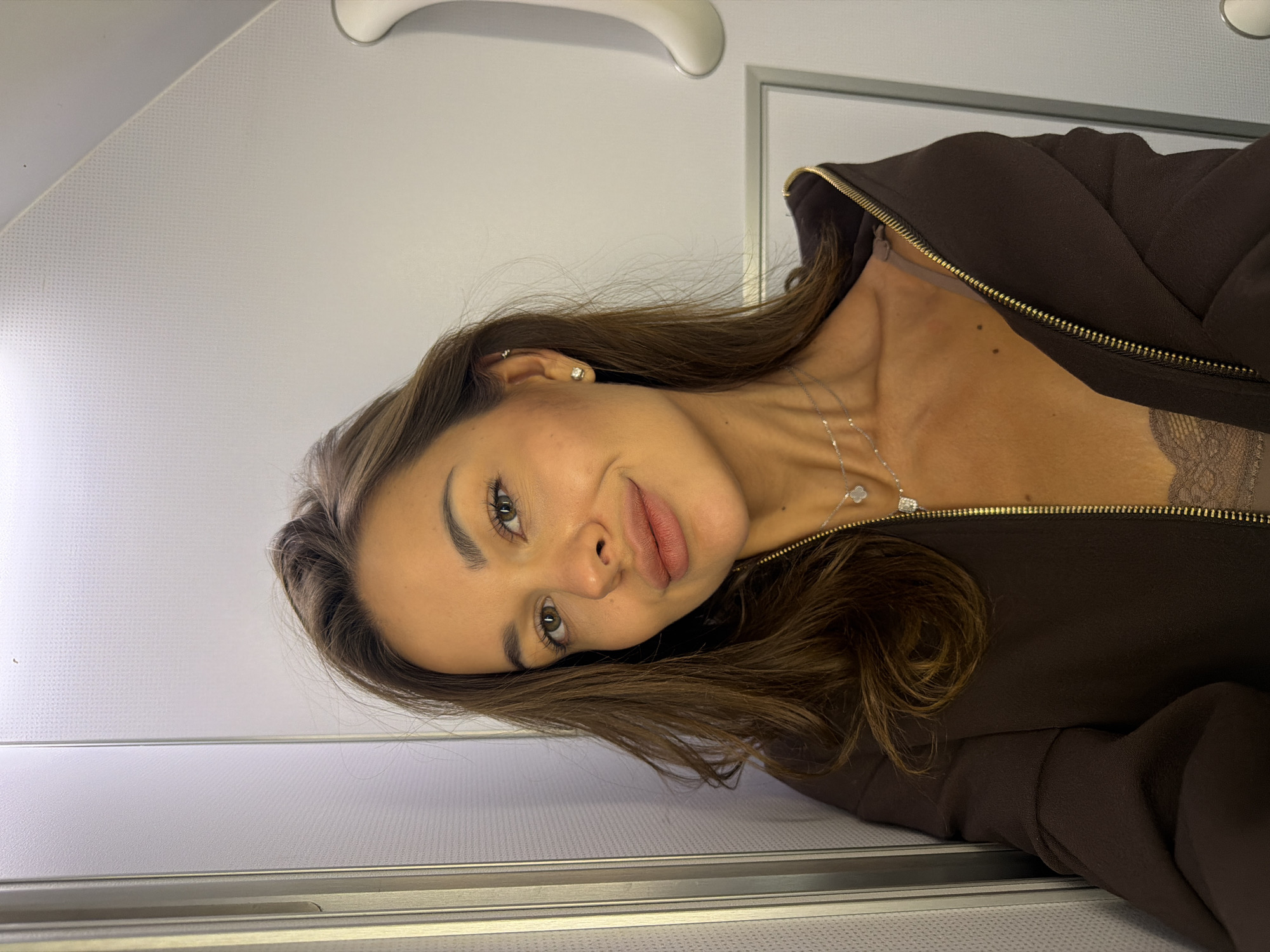 A young Caucasian woman with long brown hair is pictured in a selfie taken in a narrow, brightly lit corridor, possibly on an airplane or in a similar enclosed space. The walls appear to be paneled, with vertical metallic accents on the left and a white door or panel with a handle on the right. She is wearing a dark brown zip-up jacket, partially open, revealing a lacy brown top and a delicate silver necklace with a clover-shaped pendant. Another thinner silver chain is visible around her neck as well. She has light brown eyes and her makeup includes mascara and lipstick. The lighting is overhead and bright, casting a slight shadow on her face. The background suggests a confined, utilitarian environment. There is no visible indication of the city of Перепечино, Russia.