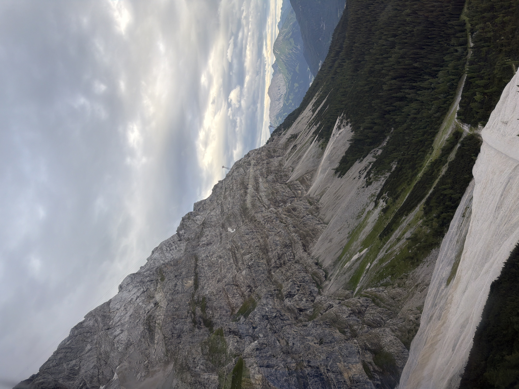 The image depicts a dramatic, rocky mountain slope on the Zugspitze, Germany's highest peak, located near Grainau. A steep, grey, and weathered cliff face dominates the left side of the frame, with visible layers of rock and patches of green vegetation clinging to its surface. A thin cable, likely part of a cable car system, is stretched diagonally across the upper portion of the mountain.
The slope transitions from the sheer rock face to a gentler, scree-covered incline dotted with sparse green grass. Further down, a dense, dark green coniferous forest blankets the lower slopes, creating a stark contrast with the lighter mountain above. The forest floor appears uneven, with a visible track or path winding through the trees.
In the distance, rolling hills and subsequent mountain ranges are visible, receding into a hazy, atmospheric perspective. The sky is overcast with heavy, grey clouds, suggesting a cool or potentially wet day. No people or specific actions are depicted in the scene. The overall impression is one of rugged, natural grandeur and the imposing scale of the alpine environment.