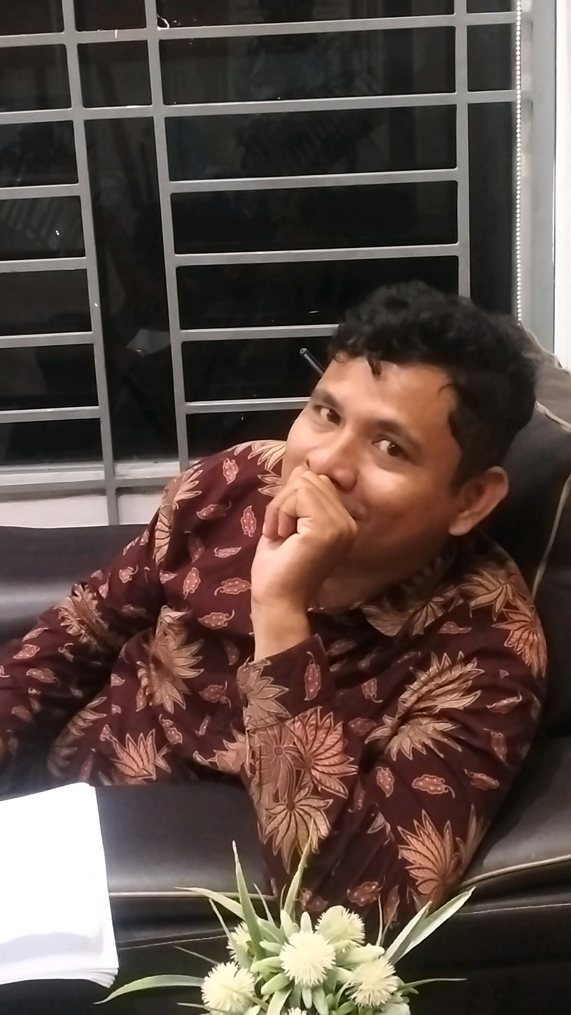 A medium shot photograph shows a man with dark curly hair and medium-toned skin sitting on a dark brown leather couch. He is wearing a maroon batik shirt with a floral pattern in tan and cream. The man is leaning his right elbow on the couch and has his right hand to his face, with his fingers touching his mouth. He is looking towards the viewer with a slight smile. Behind him is a dark window with a gray metal grate. In the foreground, on the left side of the image, is a stack of white paper. To the right of the paper is a cluster of light green artificial flowers. The overall lighting is dim, suggesting an indoor setting, possibly at night. The image is consistent with a photograph taken in an indoor residential or office setting within the city of Lhokseumawe, Indonesia, given the characteristic batik attire and common interior design elements.