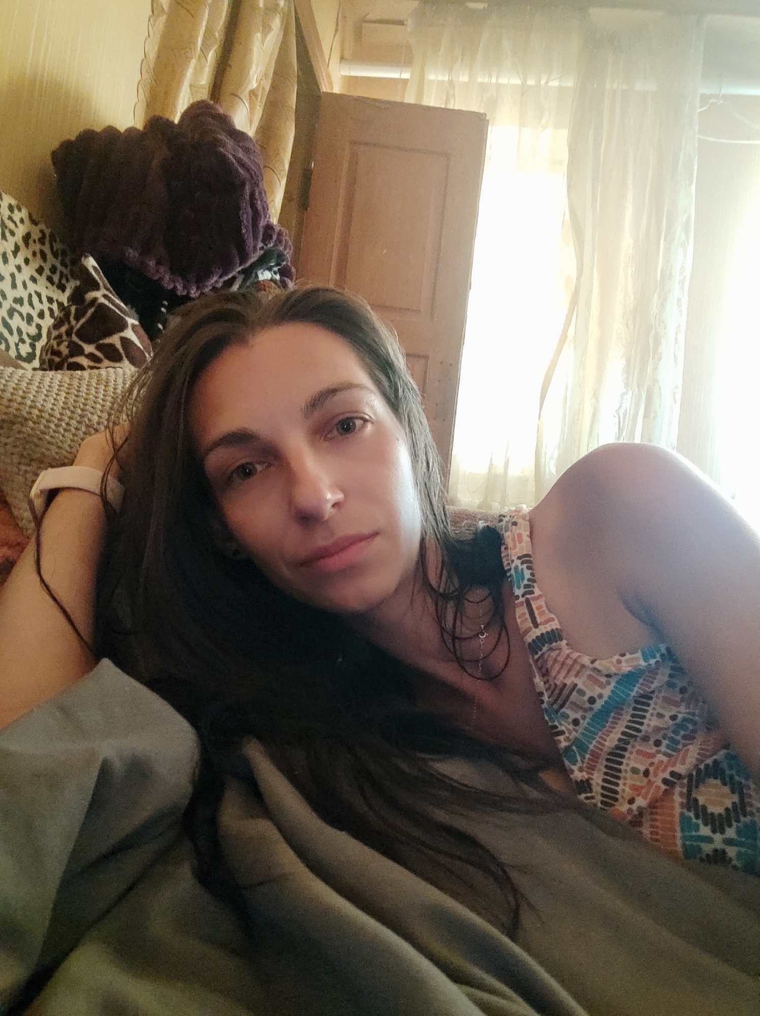 The image is a close-up, indoor, selfie-style photograph of a woman lying on a bed. The woman has long, dark hair and is wearing a colorful, patterned top. She is looking directly at the camera with a relaxed expression. The background shows a dimly lit room with curtains and a wooden door. There are also some objects visible in the background, including what appears to be a woven basket and some folded fabric. The overall lighting suggests it is daytime, with light coming from a window behind the woman. The specific location in Kryvyi Rih, Ukraine, is not determinable from the image.
