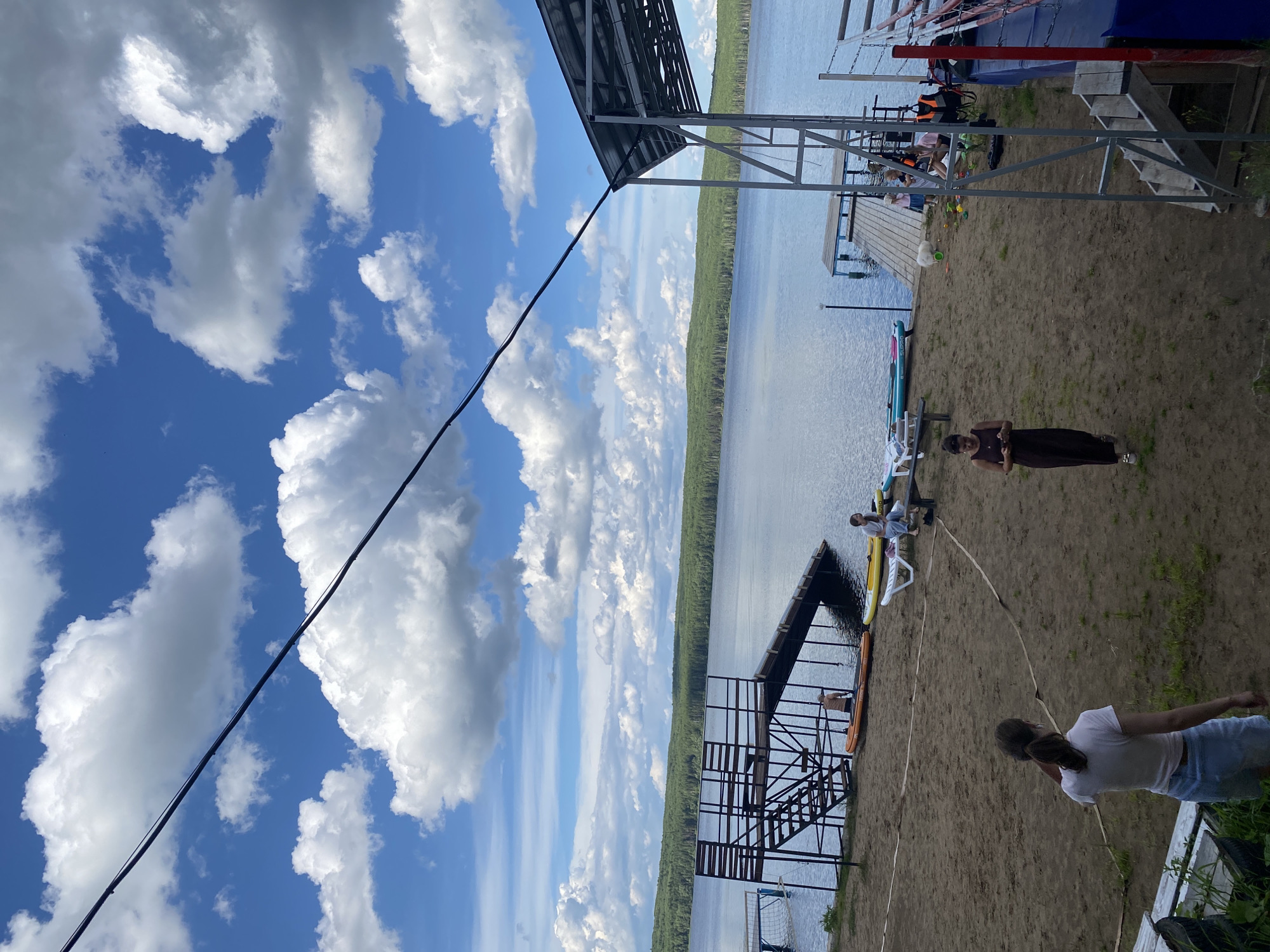 The image depicts a sandy beach area next to a calm body of water, likely a lake or river, with a dense green forest bordering the opposite shore. The sky is a vibrant blue with scattered white, puffy clouds, suggesting fair weather. A metal structure with a roof, possibly a pier or a covered seating area, extends over the water. A dark cable runs diagonally across the frame, from the lower left towards the upper right.
Several people are visible on the beach and near the water. In the foreground, a person in a white t-shirt and blue shorts is looking away from the camera, their back to the viewer. Further back, closer to the water, another person in a dark dress is standing. A few other individuals are scattered around, some sitting on a pier or on lounge chairs. Two kayaks or paddleboards are visible on the sand near the water. A metal staircase leads down to a lower platform from the covered structure.
The scene appears to be a recreational area, possibly a resort or a public beach, where people are enjoying a day outdoors by the water. The overall impression is one of a relaxed summer day. The lighting suggests it's daytime, likely late afternoon or early evening given the angle of the sun and the overall ambiance. There is no visible text in the image.
