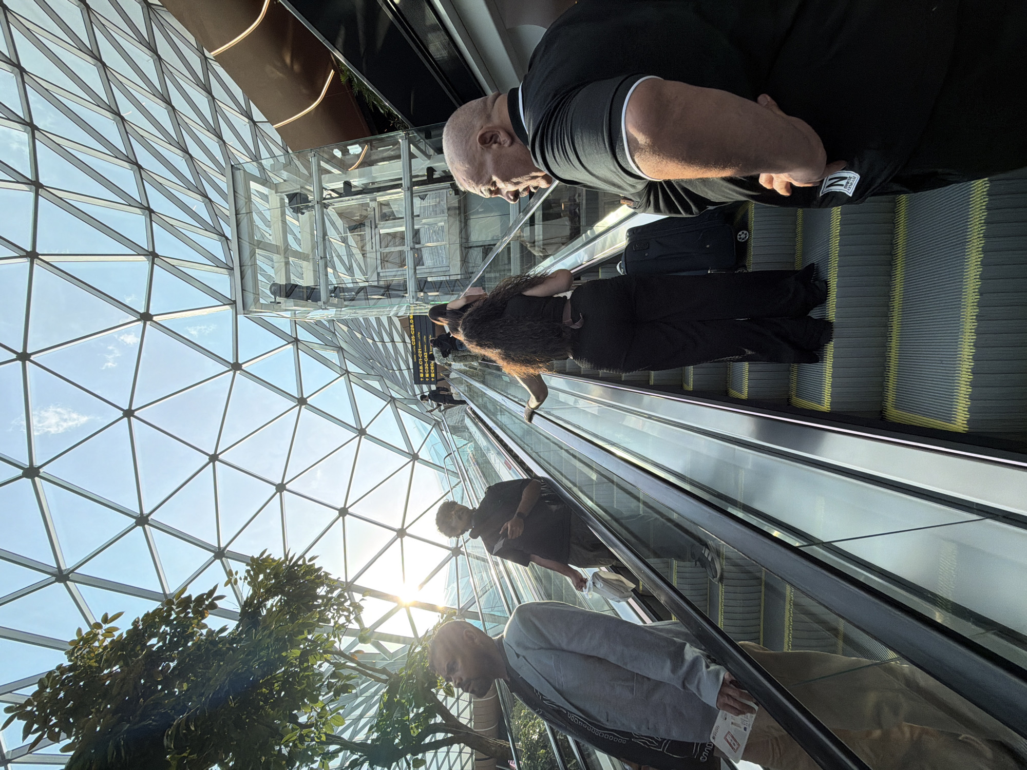 The image depicts a view looking up at a geodesic dome-like structure made of glass and metal framing, with sunlight streaming through. Several escalators are visible, with people ascending them. In the foreground on the right, a bald man with a mustache, wearing a black polo shirt with white trim, stands on an escalator, his right hand on his hip. To his left, on the same escalator, a woman with long, dark curly hair is ascending, wearing black pants and a black top. She has a black suitcase with her. On a parallel escalator to the left, a man in a grey blazer and khaki pants is ascending, holding a brochure or paper in his left hand. Further up the escalator, another man in a black t-shirt is ascending, holding a mobile phone and a plastic bag. The overall setting appears to be a modern indoor space, possibly an airport terminal or a large public building. This scene is located at Jewel Changi Airport in Singapore, not Doha, Qatar.