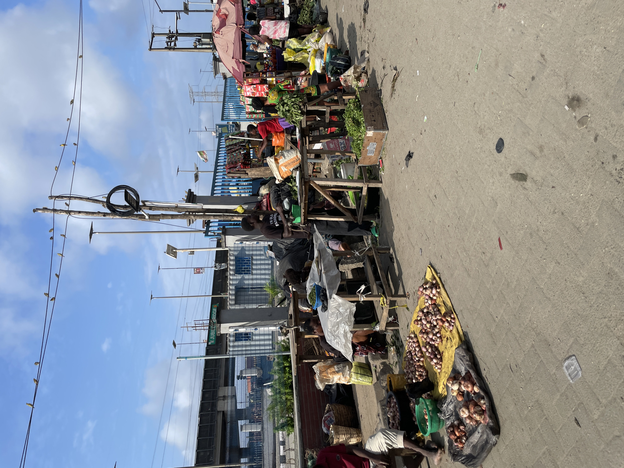 In Lekki, Nigeria, a bustling outdoor market scene unfolds under a partly cloudy sky. Several individuals are engaged in commerce, tending to stalls laden with produce. Piles of onions are prominently displayed on yellow and black tarps, with some already bagged in plastic containers. Baskets containing various goods are scattered around. In the background, a blue fence encloses what appears to be an industrial or transportation area, with a train track and overhead wires visible. Streetlights and utility poles punctuate the urban landscape. The ground is paved with concrete and shows signs of wear and scattered debris.