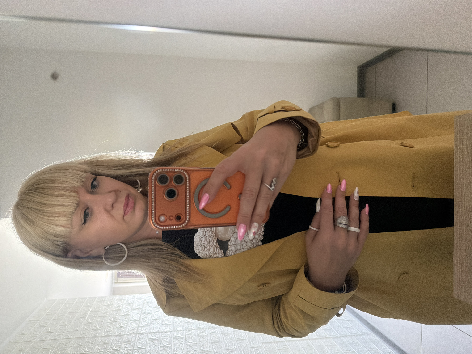 A woman with blonde hair and bangs is taking a selfie in front of a mirror. She is wearing a mustard-colored trench coat over a black top adorned with a white circular design. She's holding an orange-colored phone with a 'C' decoration on its back. Her fingernails are long and painted in pink and white designs. She is wearing large hoop earrings and rings on her fingers. The background shows a room with textured white walls and a glimpse of what appears to be a tiled floor or wall paneling. The environment suggests an indoor space, possibly a bathroom. There's no definitive information in the image to confirm the location in Chaikovsky, Russia.