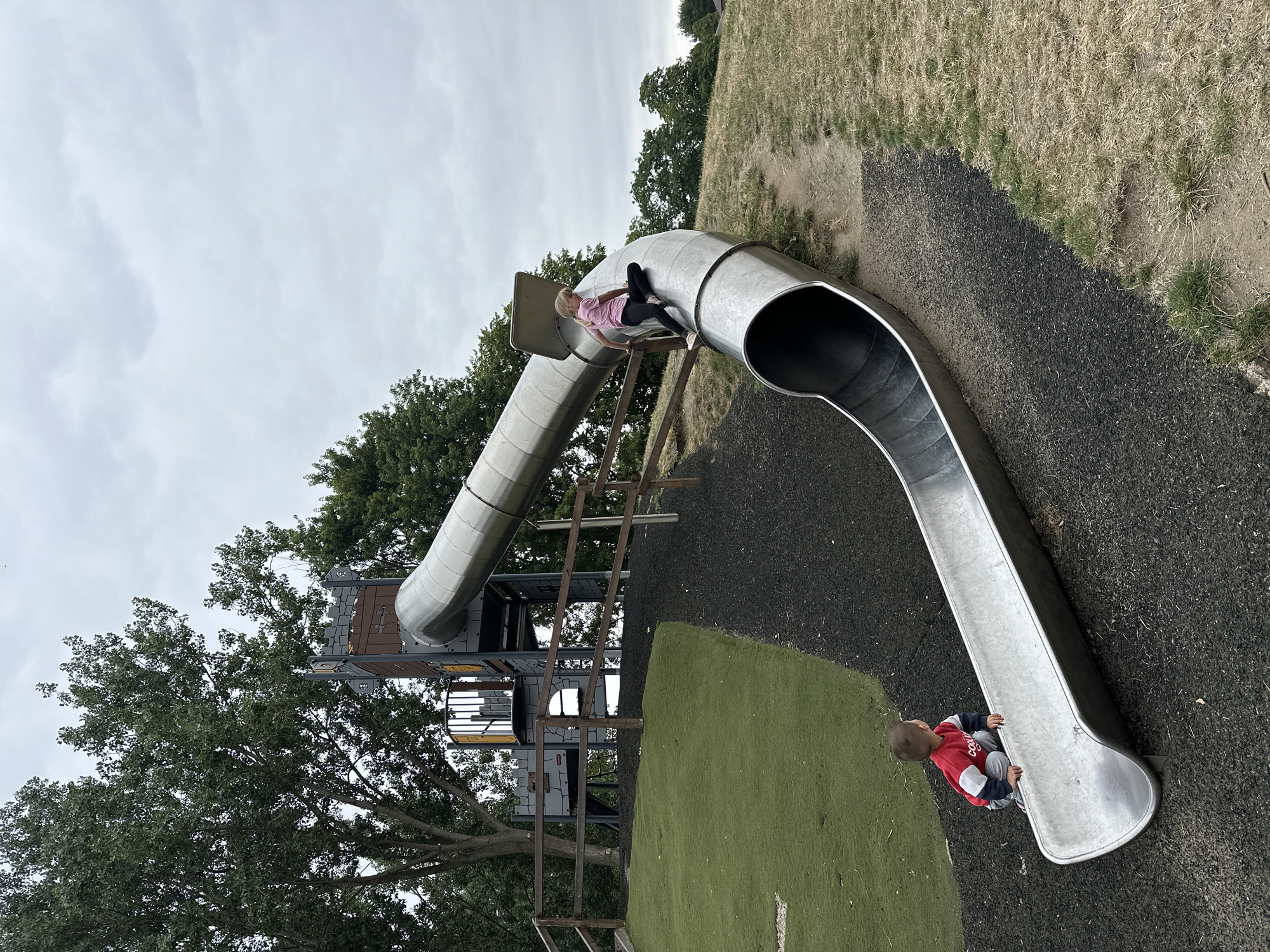The image depicts a playground scene on an overcast day. A large, curved metal slide dominates the foreground and midground, with a young child in a red, white, and blue top and grey pants sitting at the bottom of the slide. Further up the slide, a girl in a pink t-shirt and black leggings is positioned as if sliding down. Behind the slide structure, a castle-themed play area is visible, complete with turrets and walkways, adjacent to mature trees. The ground surrounding the slide consists of dark, loose material, likely rubber chippings, with patches of dry grass and a section of artificial turf on a grassy mound. A wooden fence runs along the perimeter of the mound. The location is specified as a playground in the city of Wakefield, United Kingdom.