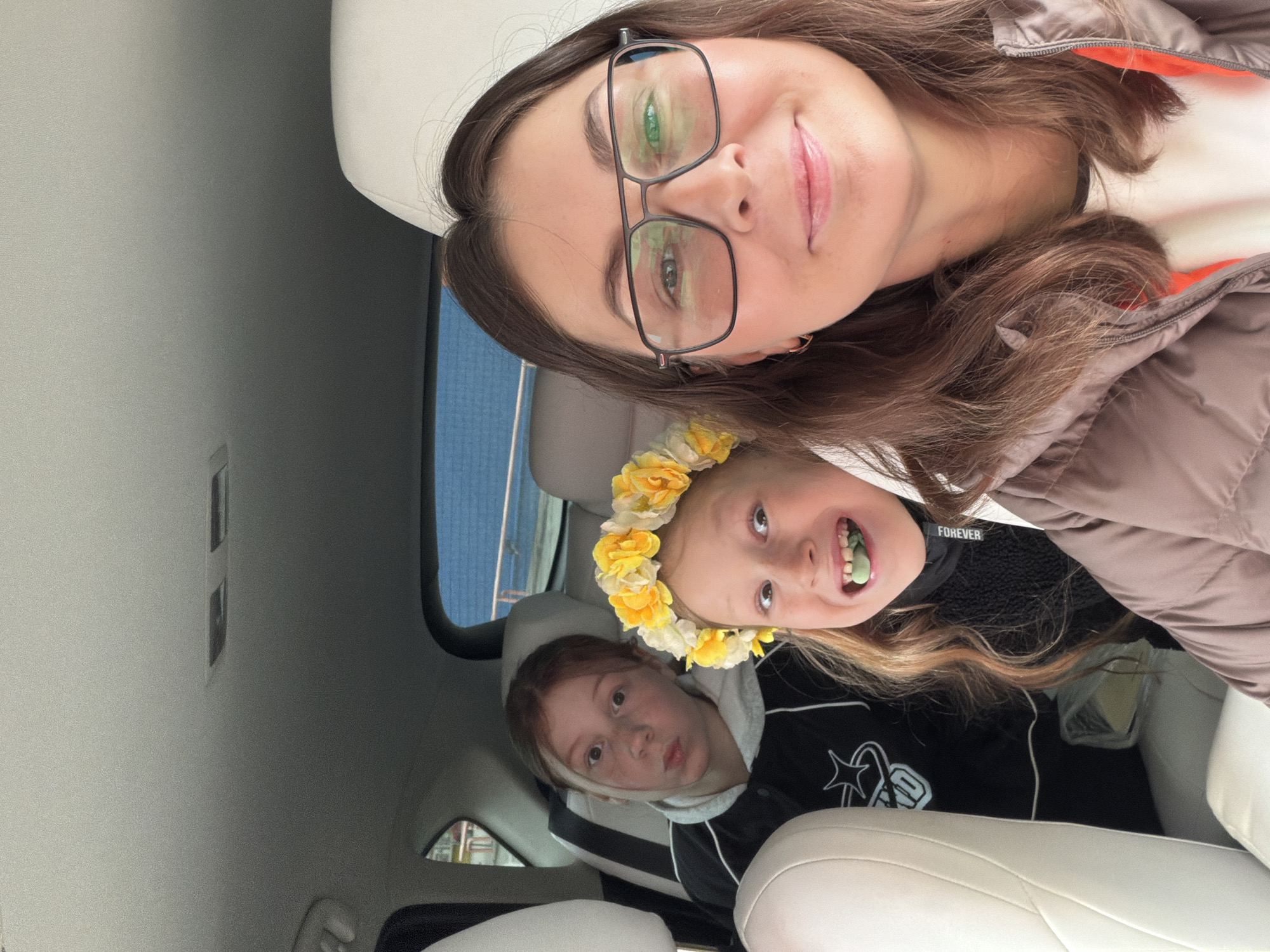 A selfie taken inside a car captures a woman and two young girls, with the image rotated 90 degrees counter-clockwise.
The main subject, a woman with long brown hair and black-framed glasses, smiles directly at the camera. She is wearing a brown or taupe puffy jacket with an orange lining visible around the collar.
In the back seats, two young girls are visible. The girl closer to the foreground has light brown or blonde hair and wears a vibrant yellow floral wreath. She is smiling broadly, with something green, possibly candy or gum, in her mouth. Her dark top clearly shows the text "FOREVER" on the collar. Next to her, another young girl with reddish-brown hair looks towards the camera with slightly pursed lips. She is dressed in a dark hoodie featuring white drawstrings and a white star-shaped design on the front.
The car's interior is characterized by light-colored (cream or white) seats and headrests. The blurred background seen through the windows suggests motion or a focus on the subjects, with hints of blue from the sky or a building. The scene is well-lit by natural light, indicating it is daytime. The overall emotion conveyed is one of casual happiness and playfulness, typical of a family moment during a car ride in a general setting, with no specific landmarks identifying Chaikovsky, Russia.