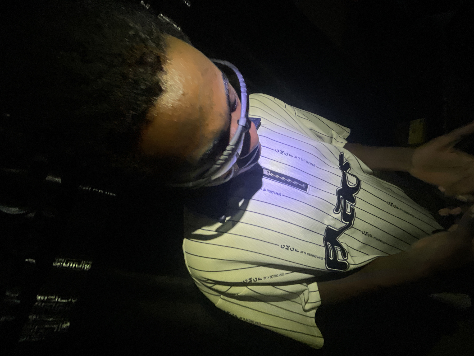 A person is seen at an angle, facing slightly away from the viewer. They are wearing a white and black striped shirt with writing on it and headphones around their neck. The environment is dark, making it difficult to discern the specifics. The location is not discernible from the image.