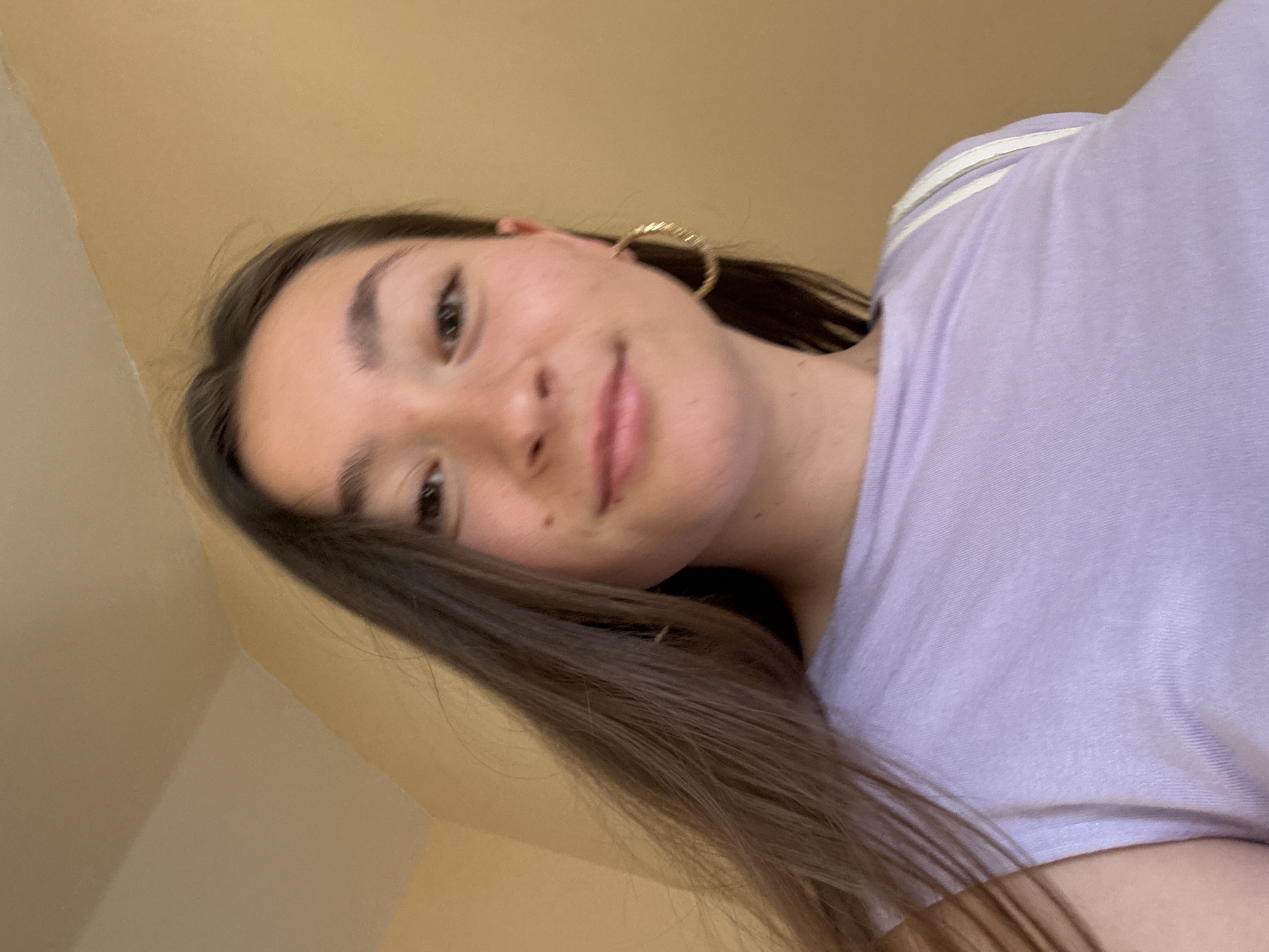 This media file is a close-up, low-angle, selfie-style photograph of a young woman with long, dark brown hair and fair skin. She is wearing a light purple t-shirt with white stripes on the shoulder area. She is also wearing a large, textured gold hoop earring in her visible left ear. Her expression is a gentle smile, and she appears relaxed. Her eyes are dark brown and she has a small beauty mark on her cheek. The background appears to be a wall with a warm, yellowish-brown hue. The lighting suggests it is daytime, likely indoors, as there are no strong shadows or indications of natural outdoor light. The orientation of the photograph is slightly tilted, as if the person is holding the camera above their head and looking down. No text is visible in the image. The location context is Sofia, Bulgaria, but there are no specific visual cues within the image to confirm this.