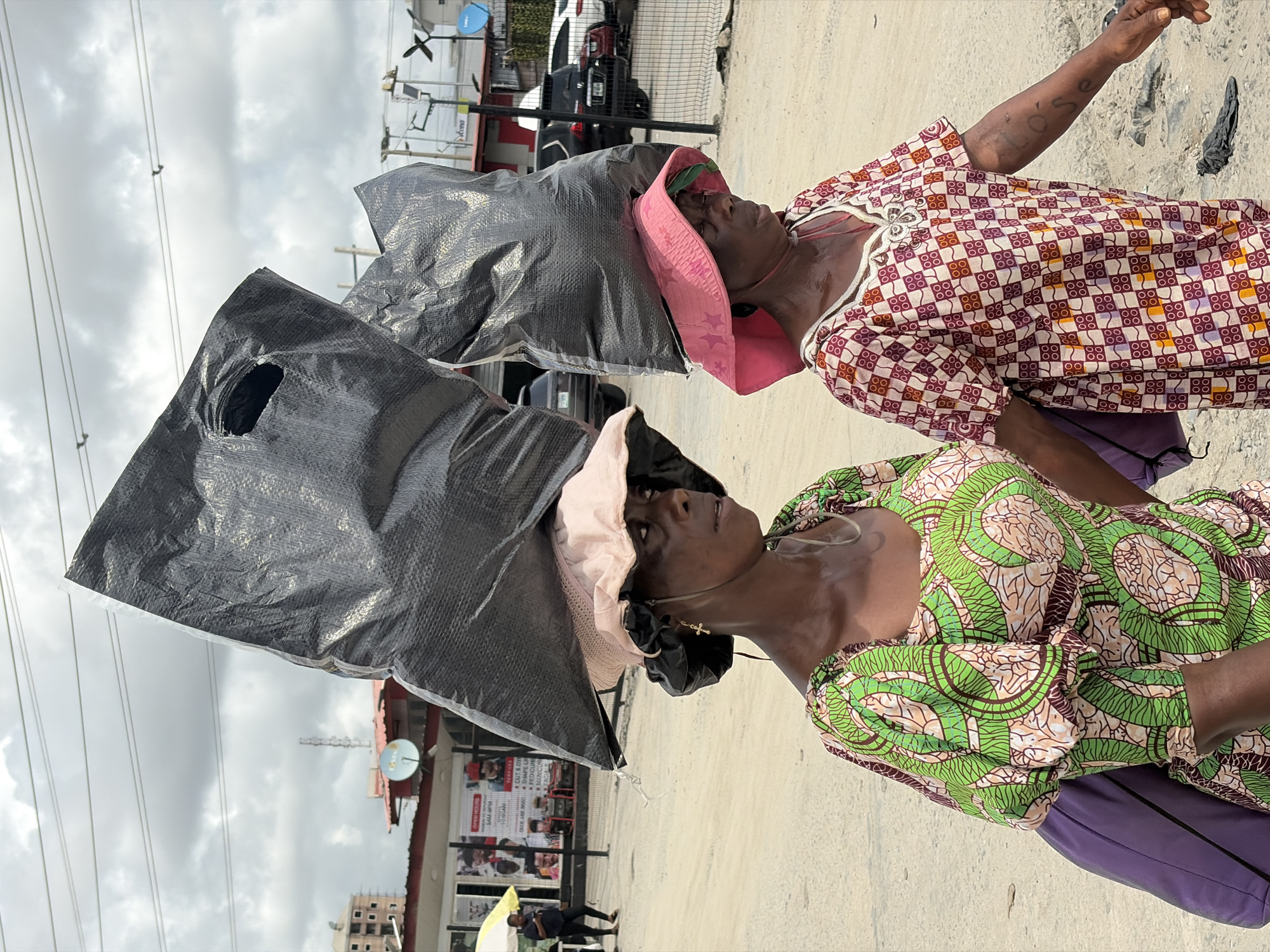 This image captures two women standing outdoors in an urban setting in Igboefon, Nigeria, likely during daytime under an overcast sky. Both women are prominently featured, carrying large, dark, possibly tarpaulin-like bags balanced on their heads, a common practice for transporting goods.
The woman in the foreground on the left wears a vibrant green, brown, and light-patterned long-sleeved dress and a light-colored hat beneath her headload. She has a small cross-shaped earring. The woman positioned behind her, slightly to the right, is dressed in a checkered reddish-brown, white, and orange patterned long-sleeved dress, with a pink hat adorned with white stars under her headload. A word, "L'ose", is visibly written on her upper right arm, possibly a tattoo or mark. Both women appear to be looking forward or slightly upwards with neutral expressions.
The background depicts a bustling street scene with power lines stretching across the sky. Buildings are visible, some featuring a blue satellite dish and what appear to be solar panels. Security fencing and various vehicles, including cars and motorcycles, are scattered in the background. Towards the left, a person is partially visible, standing under a yellow umbrella. A partially obscured sign in the mid-ground lists services such as "CUT & DRY", "SHAPE U", "PEDICURE", and "MANICURE", indicating a beauty or personal care establishment nearby. The ground is a light-colored, likely paved or dirt surface.
