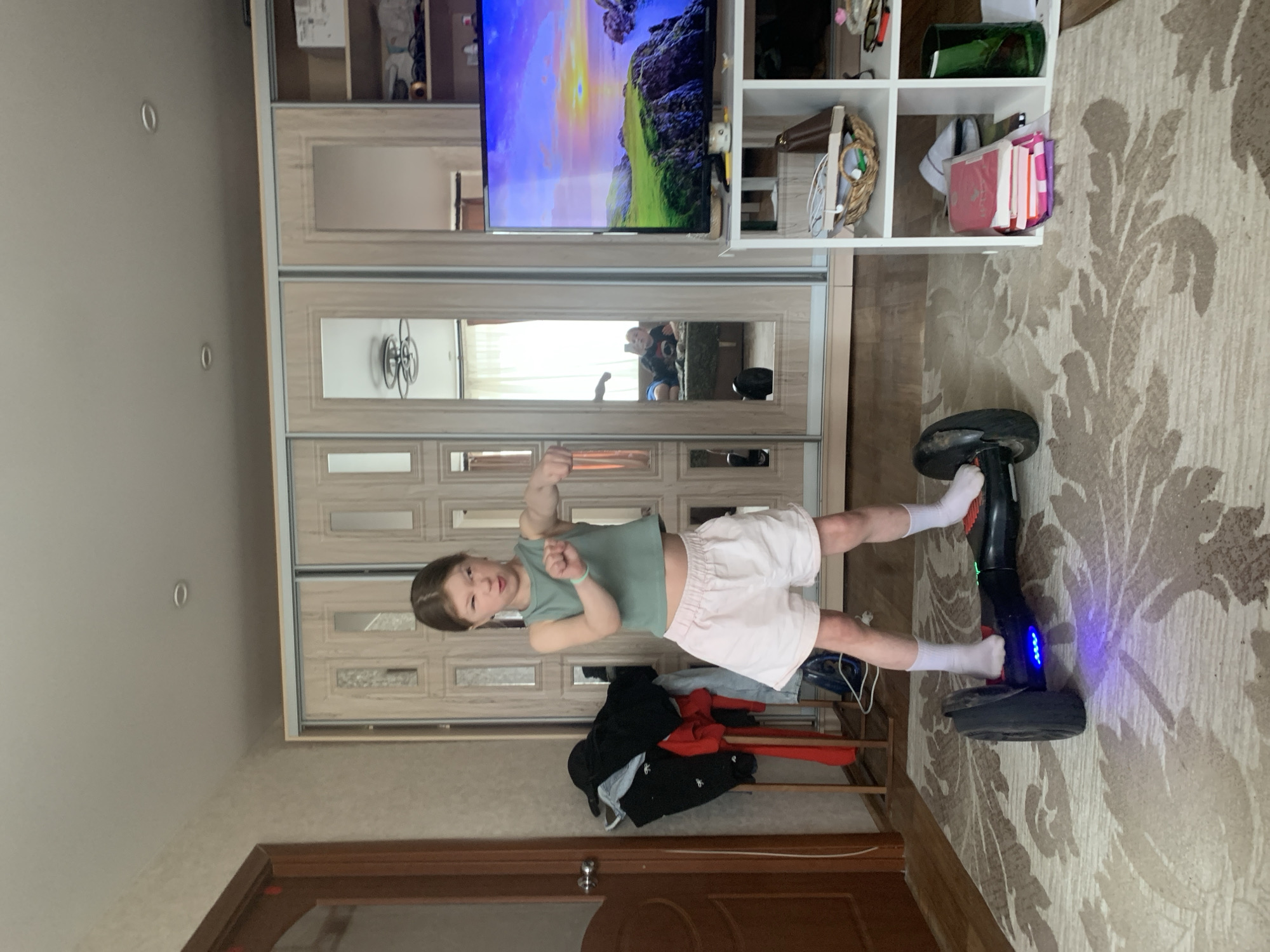 A young child stands confidently on a black hoverboard indoors, striking a pose with a slightly determined expression and a flexed arm. The child, likely a girl, wears a mint green sleeveless top, light pink shorts, and white socks. The hoverboard has blue LED lights illuminated on its front.
The setting appears to be a room within a home in Chaikovsky, Russia, characterized by a large, light-colored wooden wardrobe with mirrored doors spanning the background. The mirrors reflect parts of the room, including windows suggesting daylight, and another person partially visible. Above the wardrobe, a wall-mounted flat-screen television displays a vibrant animated landscape. To the right, a white shelving unit holds various household items like baskets and books.
The floor is covered by a patterned beige and brown rug. A pile of clothes, including black and red garments, lies on the floor near the child. A dark wooden door frame is visible in the bottom left of the image. The overall impression is one of playful activity in a domestic setting during the daytime.