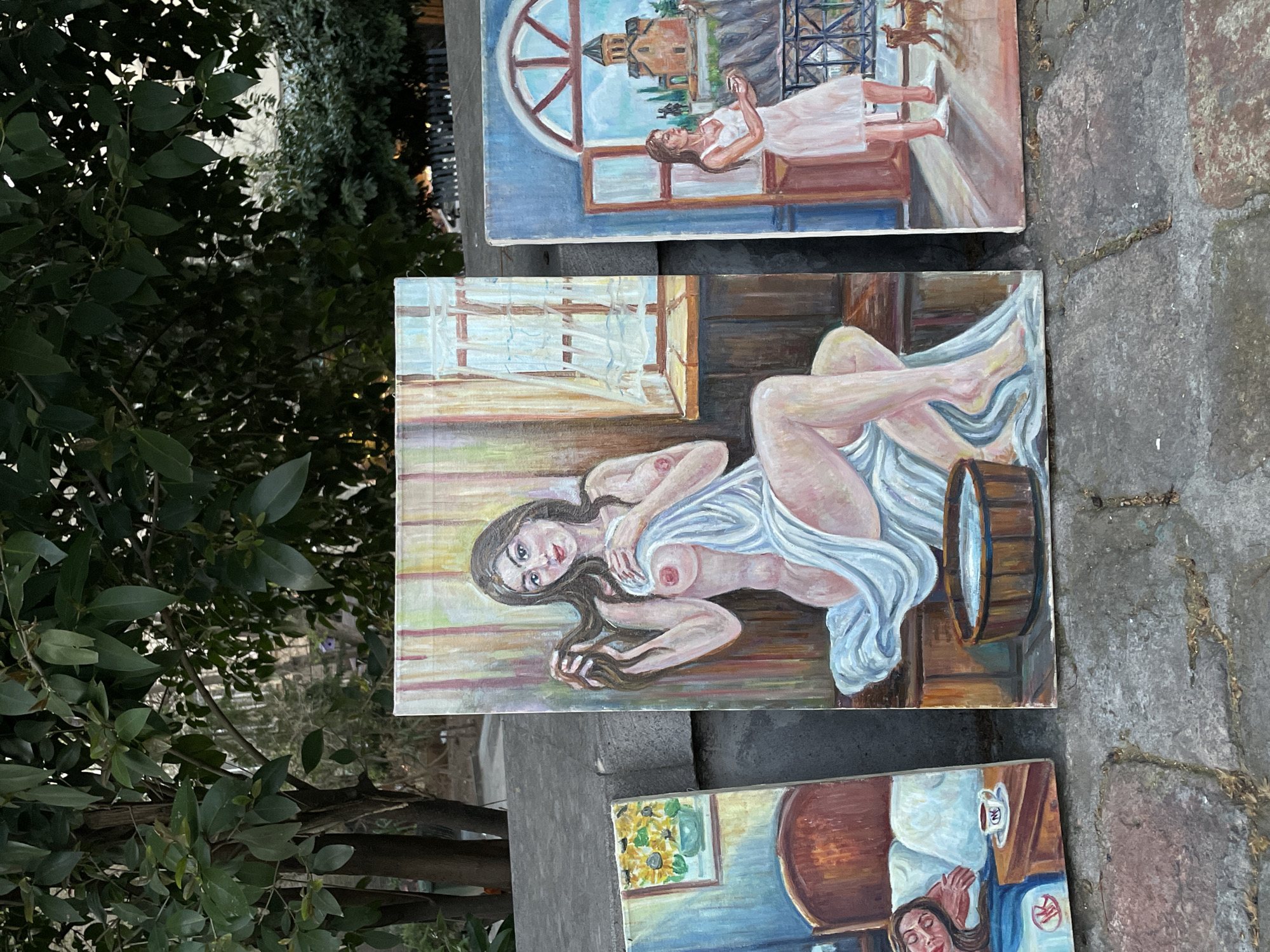 Three rectangular oil paintings are displayed outdoors, laid on a stone or concrete surface, against a backdrop of dark green foliage. The scene implies an outdoor market or exhibition in Tbilisi, Georgia, given the location context.
The topmost painting features a woman in a light dress by a large arched window, looking out at a landscape with a traditional, possibly fortified, building on a hill and a small animal in the distance. The mood is contemplative.
The middle painting shows a reclining, partially nude woman with long dark hair, covered by a white sheet, gazing directly at the viewer. A wooden tub or barrel is visible beside her. The setting appears to be an intimate indoor scene.
The bottom painting depicts a woman resting in bed, with a vase of sunflowers and a white cup on a nightstand beside her. The cup has the letters "OK" printed on it. A small red logo, possibly a stylized "K" or cross, is also visible near the bottom right of the painting. This scene conveys a peaceful, domestic atmosphere.
The overall impression is a display of art, likely for sale or viewing, under natural daylight. The diverse subjects range from serene landscapes to intimate domestic and figure studies.