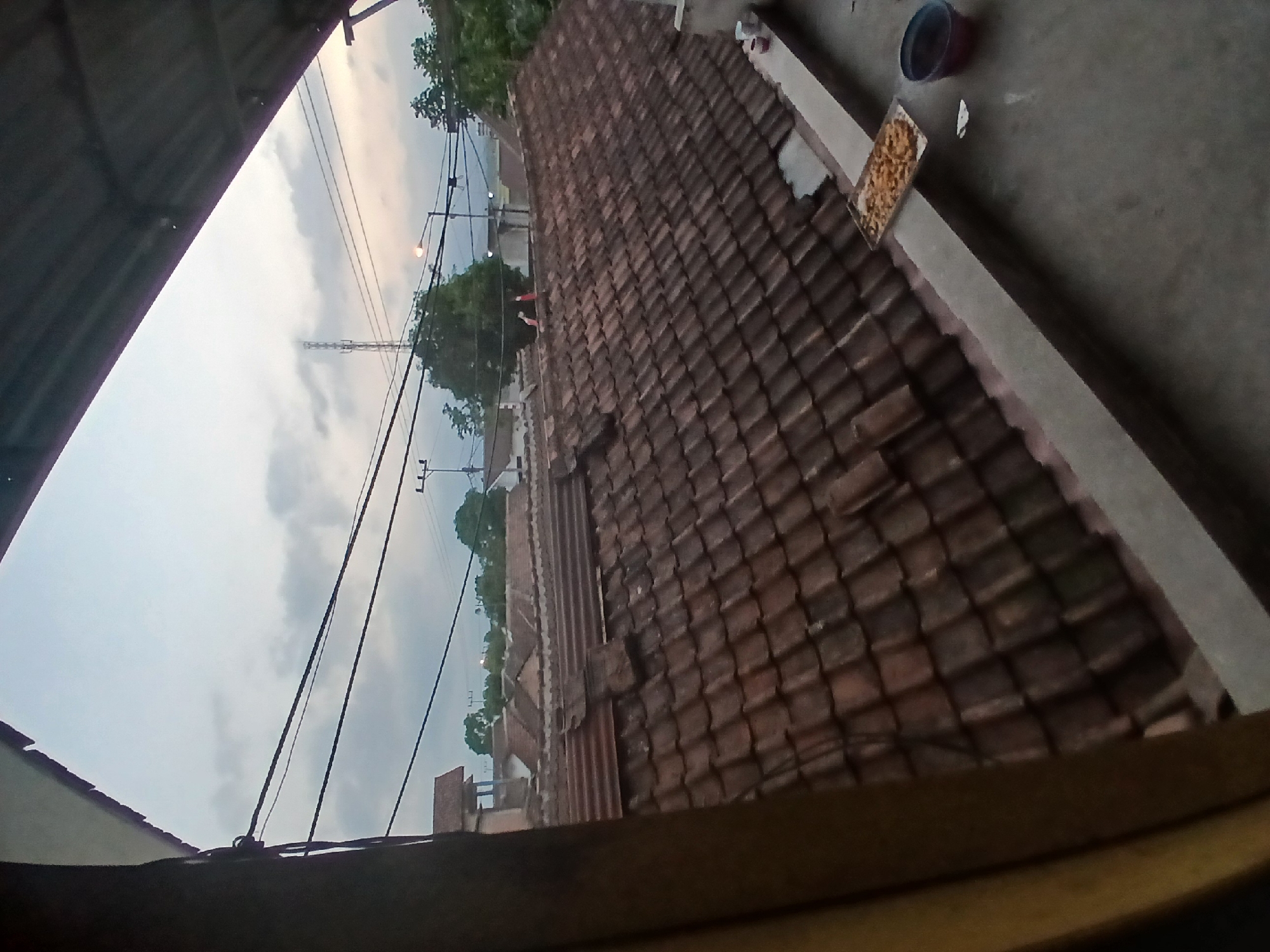 A wide-angle shot captures a view of tiled rooftops in Tenggarang, Indonesia, under a cloudy sky. Power lines stretch across the frame, intersecting with a distant radio tower. On a concrete ledge in the foreground, a tray of snacks sits next to a bucket. The scene appears to be from a residential area, with the time of day suggested to be dusk or dawn due to the ambient light.