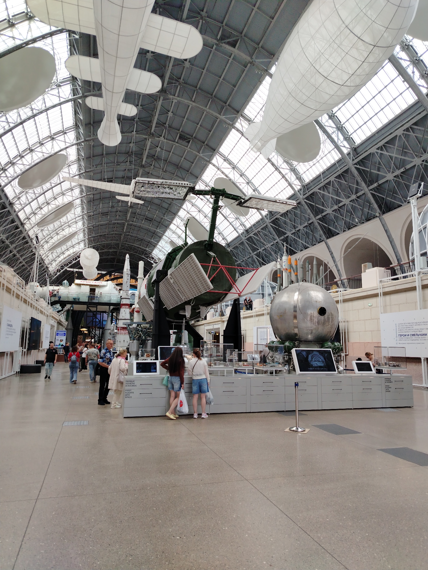 The image depicts the interior of the Memorial Museum of Cosmonautics in Moscow, Russia. The spacious hall features a high, arched, glass-paneled ceiling supported by a metal framework. Suspended from the ceiling are several white models of aircraft and airships. Dominating the central area of the exhibit are large-scale models of spacecraft, including a detailed model of a rocket and a spherical capsule with rockets attached. Several people are observed interacting with exhibits or walking through the museum. The floor is a polished concrete surface. Displays and information panels are visible along the walls and in front of the exhibits.