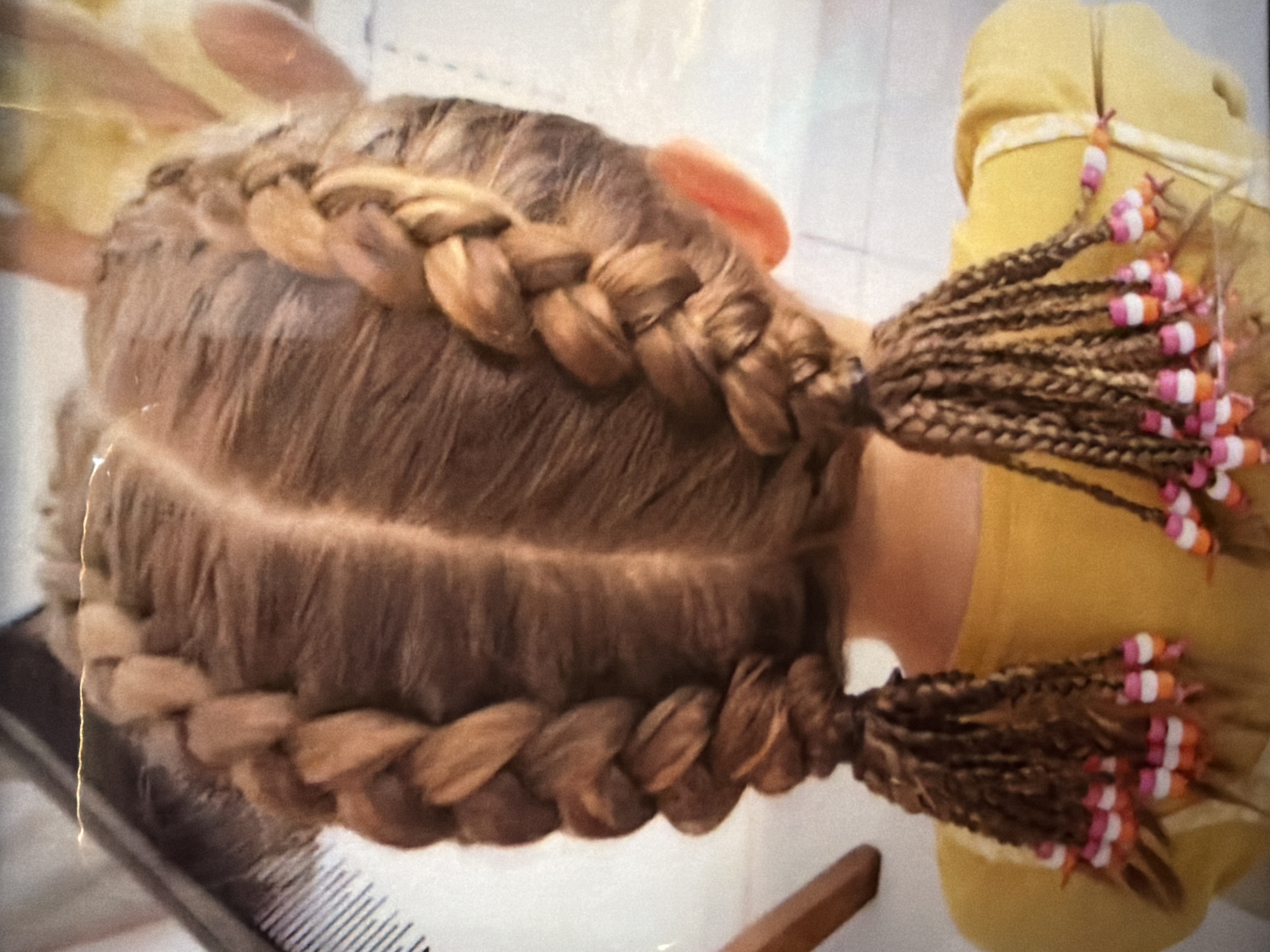 The image depicts a close-up view of a person's head with an elaborate hairstyle consisting of two thick braids that encircle the head. The braids are evenly spaced and appear to be Dutch braids. Each braid ends in a cluster of smaller, individual braids adorned with colorful beads. The person appears to be wearing a mustard yellow shirt. A portion of a comb is visible in the lower left corner. The image is taken indoors, likely in a private residence or salon.
There is no data available to indicate that the location is in the city of Kuta Selatan, Indonesia.