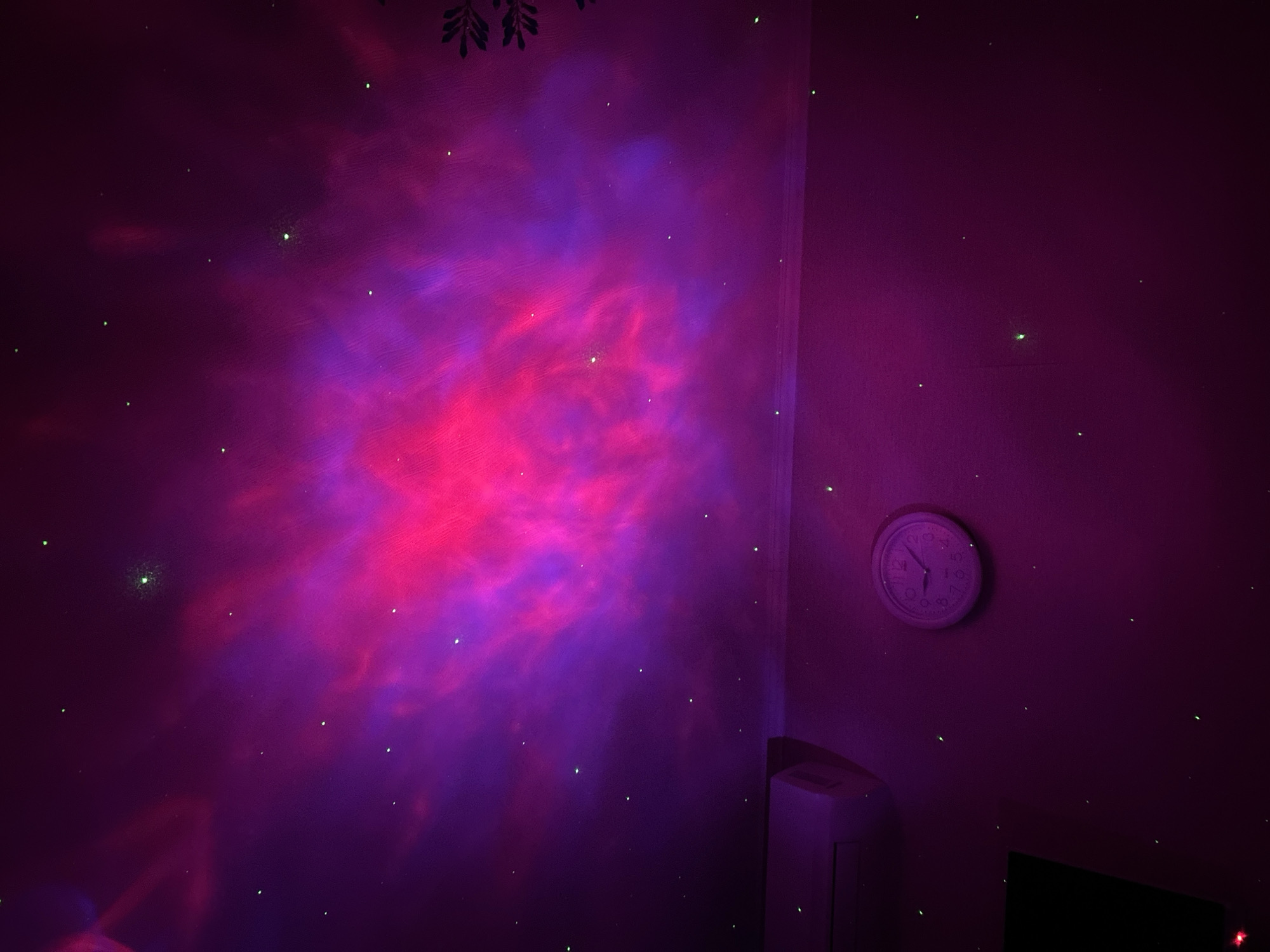 A low-light photograph depicts a room illuminated by a projection, resembling a nebula. The primary colors are shades of red, pink, and purple. The projection is cast onto a wall, where a round clock is also mounted. The clock's hands indicate a time of approximately 2:40. Green laser dots are scattered around the scene, mimicking stars. A partially visible white object, possibly an appliance or piece of furniture, is positioned near the corner of the wall. The room is located in Chaikovsky, Russia.