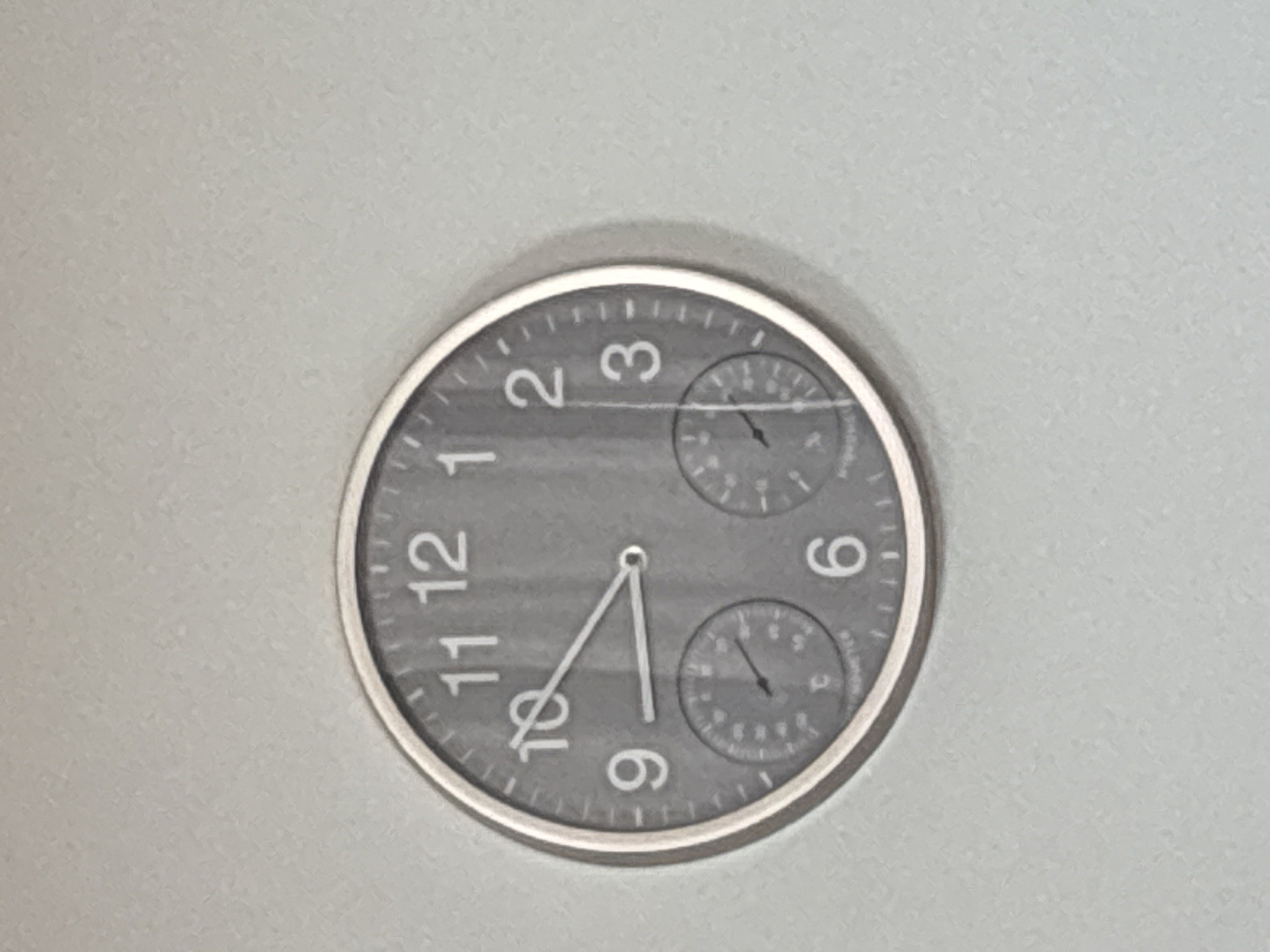 A round, grey wall clock with white numbers and hour markers is mounted on a light grey textured wall. The clock has two smaller circular dials inset at the bottom, one on the left and one on the right. The clock hands indicate approximately 9:50. The clock is the sole object in the frame. There is no identifiable location in the city of Cork, Ireland.