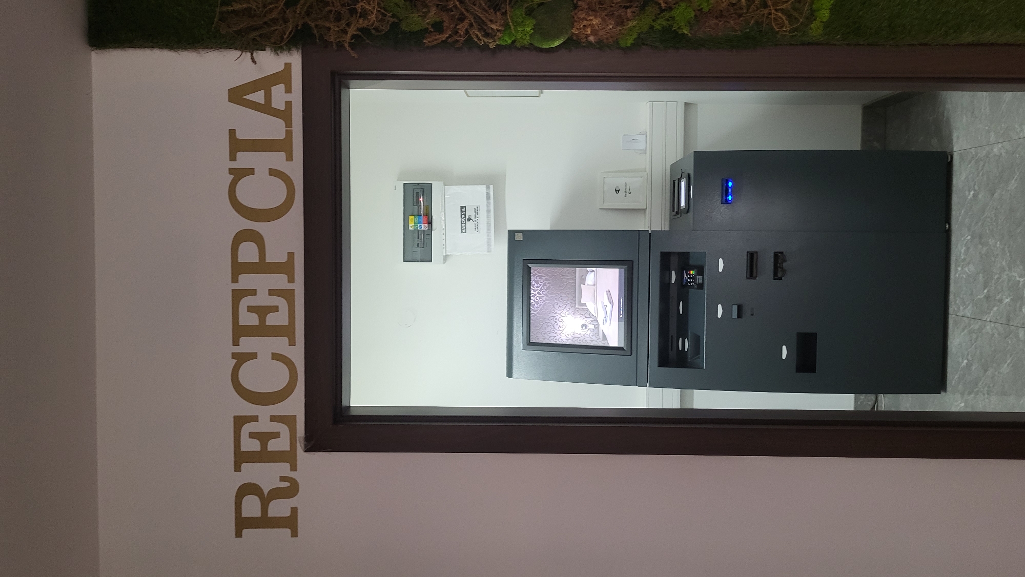A view through an open doorway reveals a dark gray ATM machine in a well-lit interior. The ATM has a screen displaying a decorative wallpaper pattern of swirling lines and a bed. Above the ATM, a white paper dispenser is mounted on the wall, and to its left is an electrical panel. To the right of the ATM, a small framed sign indicates "Wi-Fi". The wall above the doorway has large gold letters spelling "RECEPCIA", which translates to "Reception" in Slovak. The floor is tiled with a gray marbled pattern. On the far right edge of the frame, a portion of a vertical decorative panel of preserved moss and branches is visible. The overall impression is that of a reception area or lobby.