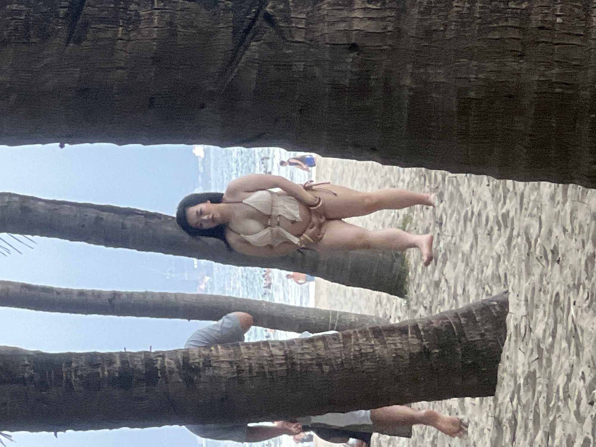 A young woman in a light-colored bikini stands on a sandy beach, partially obscured by the trunks of several palm trees. She has dark hair and is looking towards the camera. In the background, the ocean is visible with several people in the water and on the shore. The sky is overcast. The scene appears to be on a beach in the city of Đà Nẵng, Vietnam.