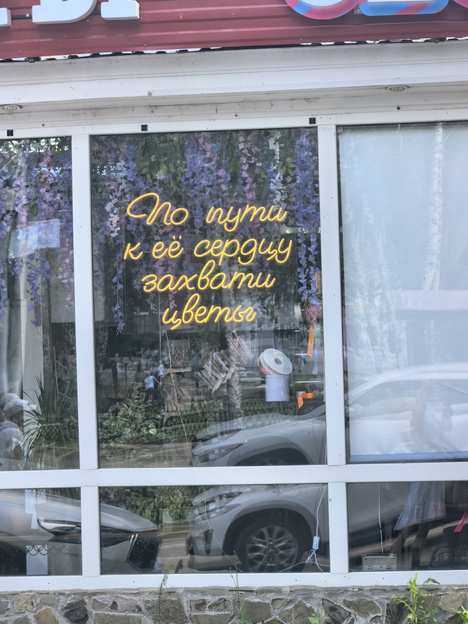 The image shows a storefront window in Sterlitamak, Russia, featuring a neon sign in Russian that reads "По пути к её сердцу — захвати цветы" which translates to "On the way to her heart – take flowers." The neon sign is yellow and illuminates a floral backdrop, possibly artificial wisteria. Inside the store, visible through the window, are plants, floral supplies like ribbon rolls, and reflections of cars outside. The storefront itself has a red corrugated metal roof and white window frames. The lower part of the building is constructed of stone.