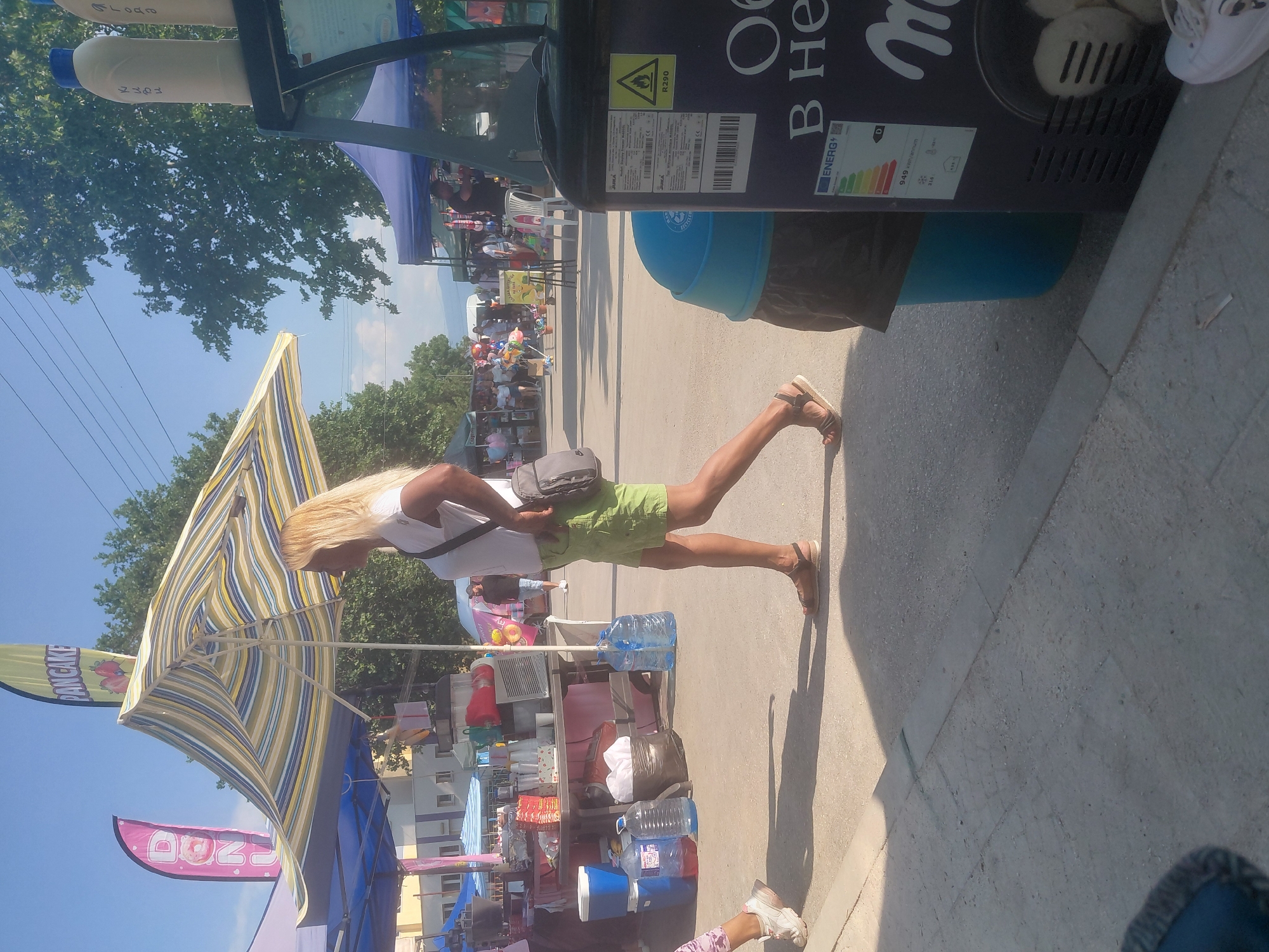 The image depicts an outdoor market or fair taking place in Kostievo, Bulgaria, under a clear blue sky. The ground consists of a paved surface, likely concrete or asphalt, showing some wear. In the foreground, slightly right of center, a woman with long, straight blonde hair walks from left to right in profile. She wears a white short-sleeved t-shirt, olive green shorts, and brown flat sandals. A grey messenger bag is slung over her left shoulder. To her immediate left, a child's left foot and lower leg are partially visible, wearing white sneakers with pink accents and light-colored patterned pants, also moving from left to right. Various market stalls and tents populate the scene. On the left, a blue-canopied tent covers a table laden with merchandise, including plastic cups, red packages, a red beverage dispenser, a silver appliance, and several large plastic water bottles. A blue cooler with a white lid and a black trash bag containing white items rest on the ground near this stall. Behind this stall, a light-colored building with windows is visible. Two prominent feather flags are displayed: a pink one on the far left reads "DONUTS" with a donut graphic, and a greenish-yellow one behind a large, open, striped (yellow, green, blue, white) market umbrella reads "PANCAKE" with fruit graphics. On the far right, a tall, dark-colored commercial refrigeration unit is visible. On its top, two cream-colored cylindrical containers with blue caps are present, marked with Cyrillic script: "КУБИ" (CUBES) and "ЯГОДА" (STRAWBERRY). The unit displays a yellow warning triangle, an "R290" refrigerant label, a barcode, and an energy efficiency label indicating "ENERGY D" and "949 kWh/annum". Inside the unit, white, round items resembling ice cream or frozen yogurt are visible. Adjacent to the refrigeration unit is a blue trash bin with a black liner. In the mid-ground and background, additional market tents and numerous indistinct people are present. One man in a dark shirt and light shorts is visible standing behind a blue tent further back on the right. Another man in a dark shirt is partially obscured behind the left stalls. Further down the street, various market goods like colorful balloons, pinwheels, and what appears to be cotton candy are visible. A white plastic chair is situated near one of the background stalls. The background features abundant green trees against the clear blue sky, with overhead power lines traversing the upper portion of