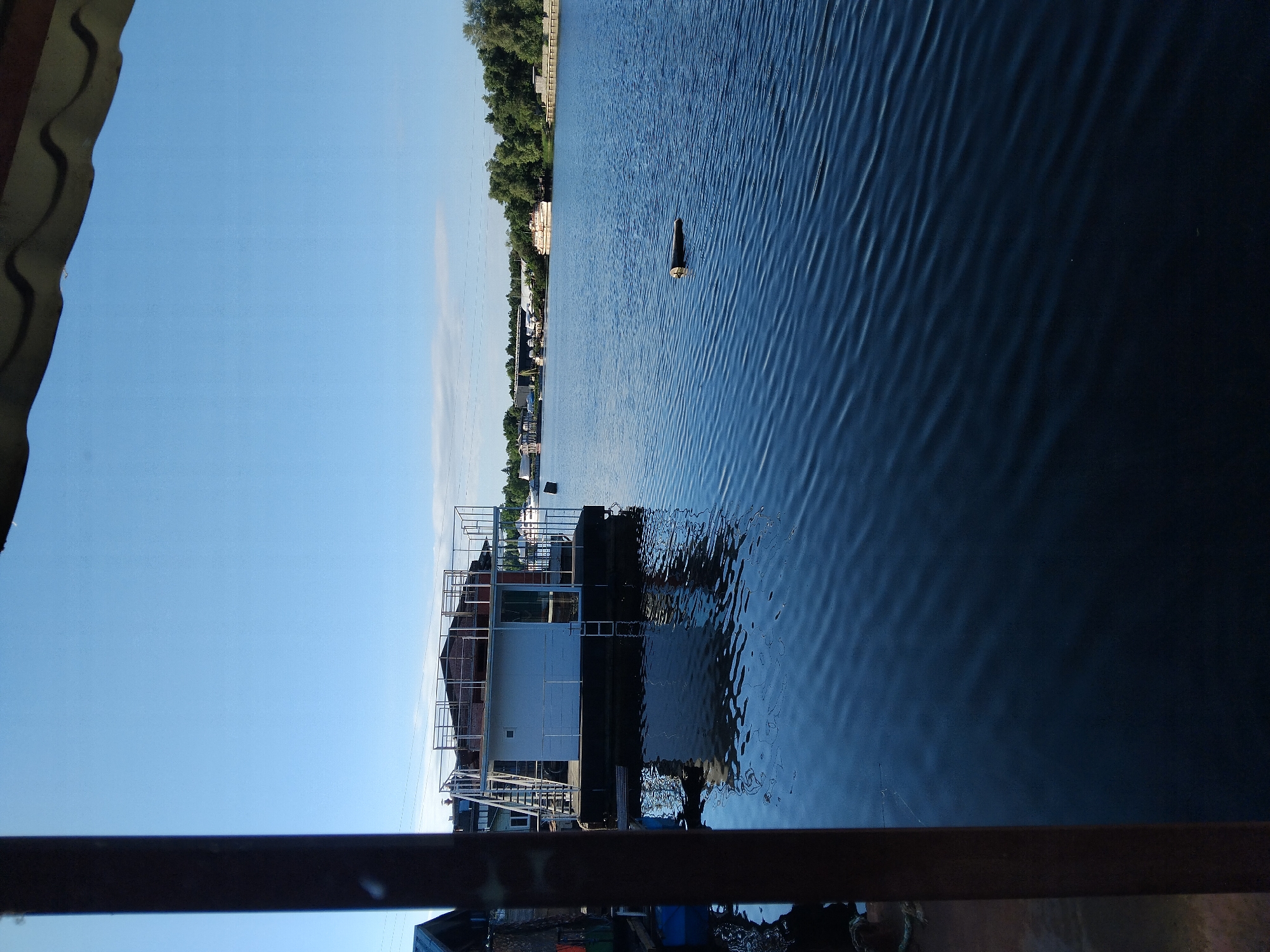 The image depicts a houseboat docked on a wide, calm body of water, likely a river, under a clear blue sky. The houseboat is white with metal railings and a ladder leading up to its roof. The water reflects the boat and the sky, creating a serene scene. In the background, there are trees, buildings, and other boats, suggesting a populated area. The perspective is from a dock or another boat, with a dark vertical pole in the foreground partially obscuring the view. This scene is consistent with the Dnipro River in Kyiv, Ukraine, which is known for its numerous houseboats and scenic riverbanks.