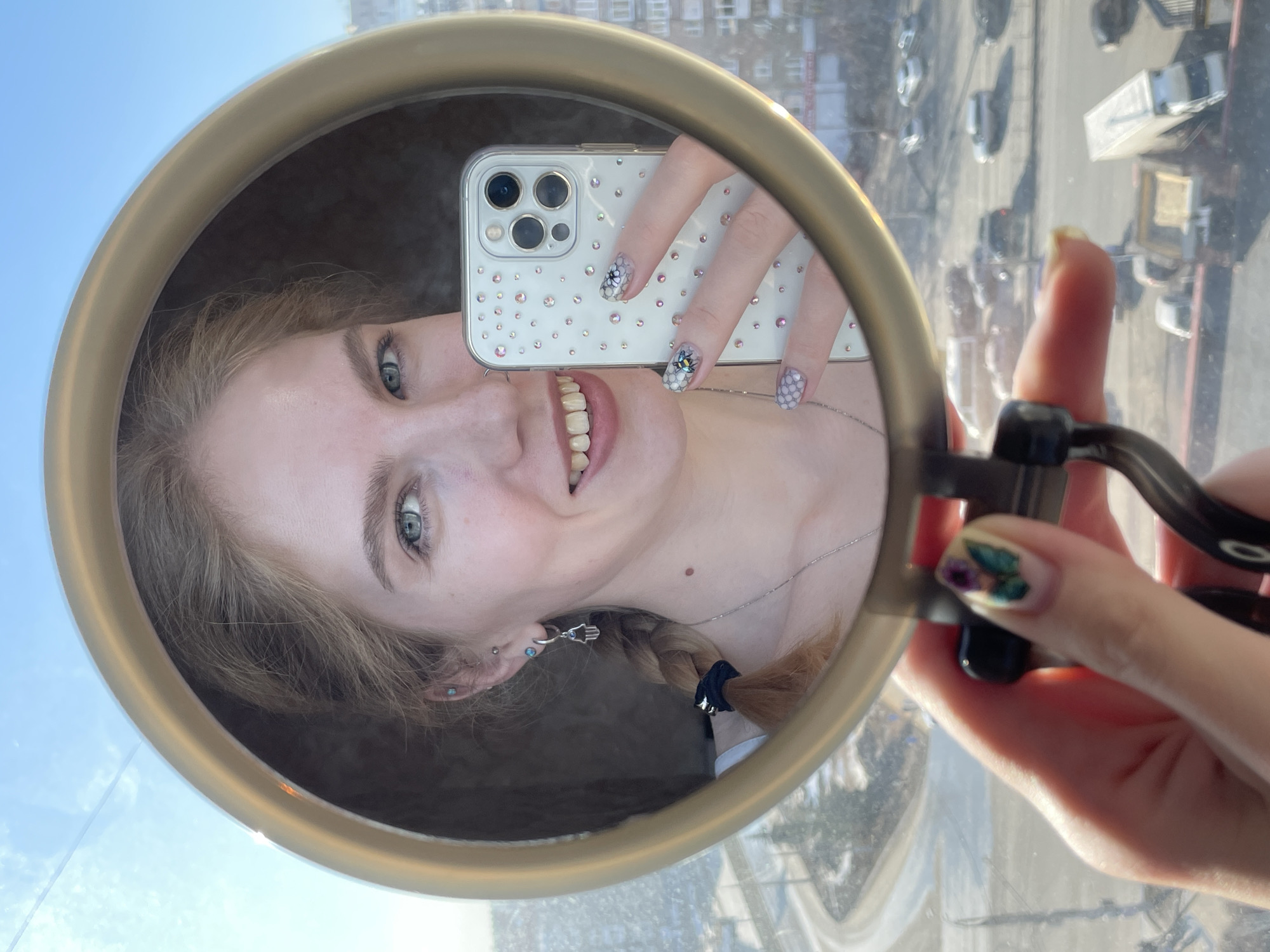 A young, fair-skinned woman is holding a round mirror with a gold frame in front of her. The mirror reflects her face, her right hand holding a smartphone with a white case and rhinestone embellishments, and the background behind her. She is smiling, and her teeth are visible. She has light blue eyes, blonde hair, and is wearing a necklace and earrings. Her right hand is visible holding the handle of the mirror and has a nail design featuring floral elements.
The background visible through the mirror and the glass panel behind it reveals a city scene, including buildings, roads, and vehicles. Based on the architecture and vehicles, and the context provided, the location appears to be Izhevsk, Russia. The weather appears to be sunny and clear.