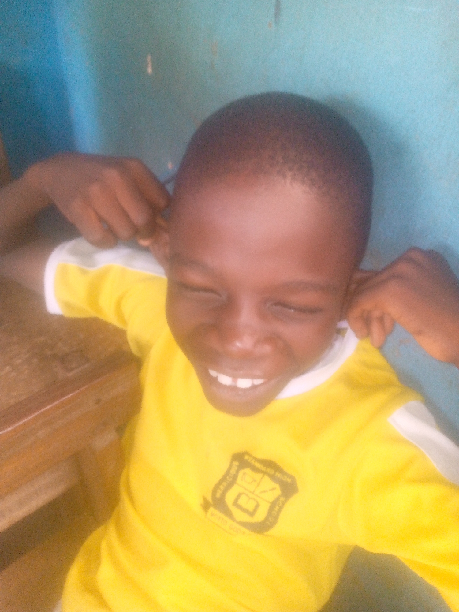 A young boy with dark skin and a close-cropped haircut is sitting at a wooden table, wearing a yellow shirt with white sleeves and a school logo on the chest. He is pulling on his ears with both hands and smiling with his eyes closed. A blue wall is visible in the background. The location is in the city of Unknown, Unknown.