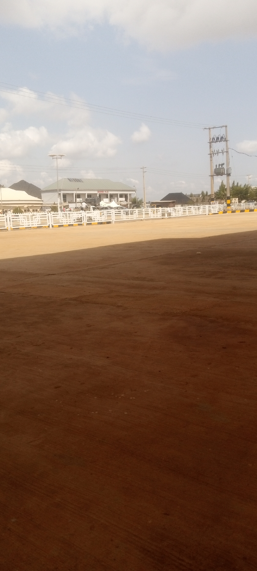 The scene depicts an open space in Sabon Tasha, Nigeria. A white, low fence with yellow accents runs horizontally across the mid-ground, partially obscuring a one-story building labeled "ALZONE PLAZA." An electrical transformer and poles stand to the right of the plaza. The foreground is a vast expanse of brown, possibly dirt or paved surface, with the lower portion appearing shaded. The sky is visible with some cloud cover.