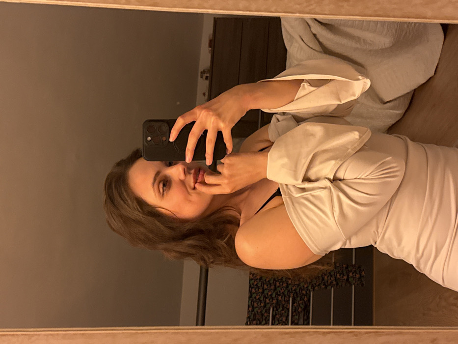 An image captured as a mirror selfie displays a female subject indoors in Cava de' Tirreni, Italy. The subject, possessing long, wavy brown hair, is positioned from the chest up, angled to her left. She is attired in a light-colored, off-the-shoulder top or dress, with a black bra strap visible on her left shoulder. Her left hand holds a dark-colored smartphone, angled to take the photograph. Her right hand is placed near her mouth, with fingertips touching her lower lip. Her expression features a subtle smile, and her gaze is directed towards the left. The reflected background reveals an interior space with warm ambient lighting. Visible elements include a clothing rack displaying dark, patterned fabric on the left, and a bed with a light-colored blanket and a dark wooden dresser on the right. The mirror itself is bordered by a light-toned frame.