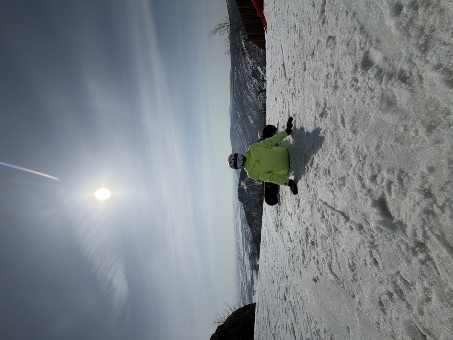 The image displays a solitary individual seated on a snow-covered mountain slope in Zelenaya Polyana, Russia, under a bright, sunlit sky. The person is viewed from behind, dressed in a neon green jacket, dark lower garments, black gloves, and a white helmet with goggles. A black snowboard is positioned on the snow directly behind the individual. Their arms are extended to the sides, with hands resting palms down on the snow. A distinct shadow of the person is cast forward onto the textured snow surface. The background features a panoramic view of snow-dusted mountain ranges and valleys under a light blue sky with faint cirrus clouds. A dark rock formation and bare tree branches are visible on the left side of the frame, while a wooden fence and a partial red object are present on the far right. A diagonal multicolored lens flare is visible in the upper right sky quadrant, originating near the sun.