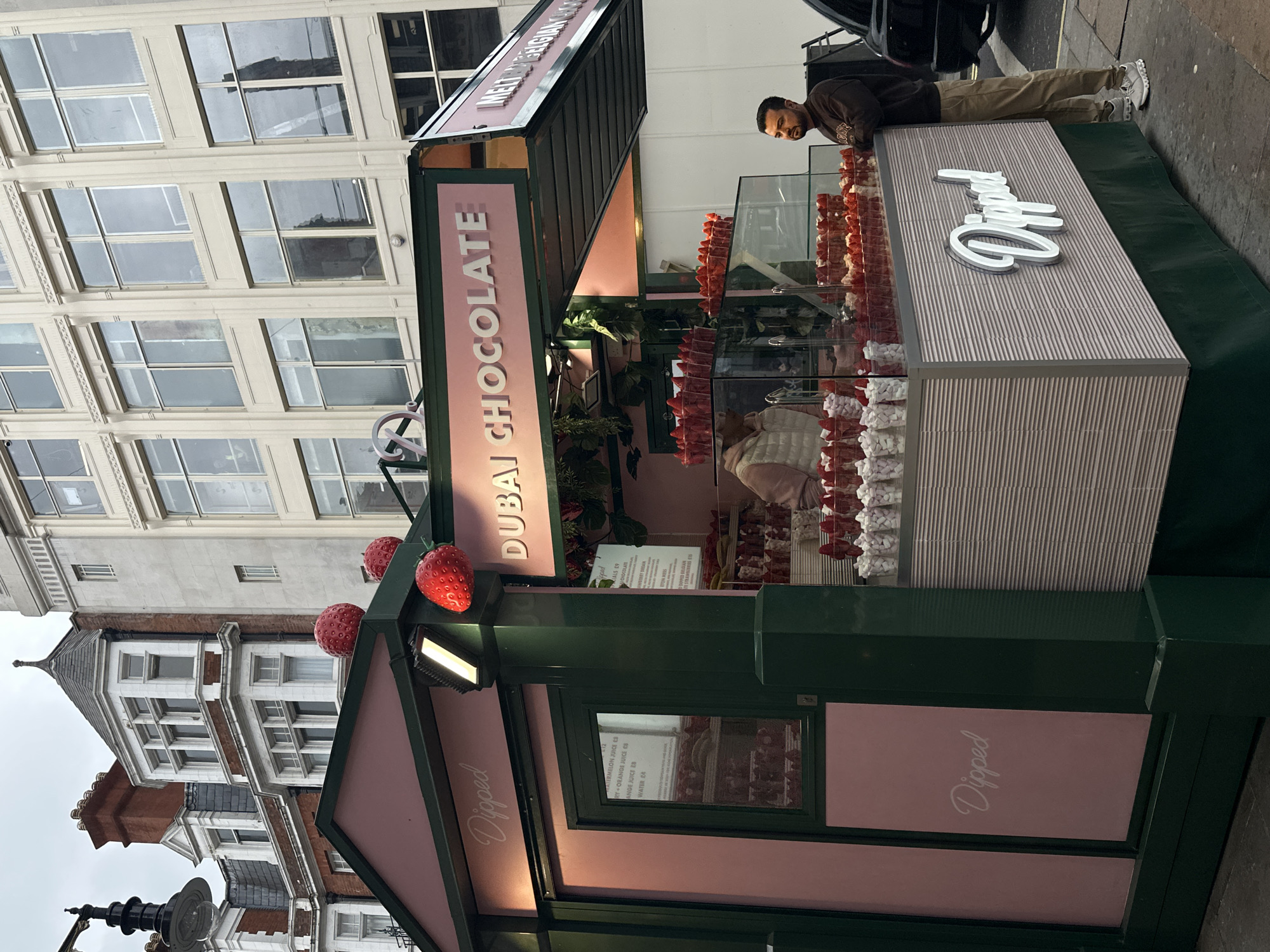 The image shows a "Dipped Dubai Chocolate" street vendor stall in London, United Kingdom. The stall is painted in green and pink, with large strawberry decorations on top. A man is standing behind the counter, possibly a vendor. The stall displays chocolate-dipped strawberries and other treats. Menus with prices are visible inside the stall. The buildings in the background suggest an urban setting.