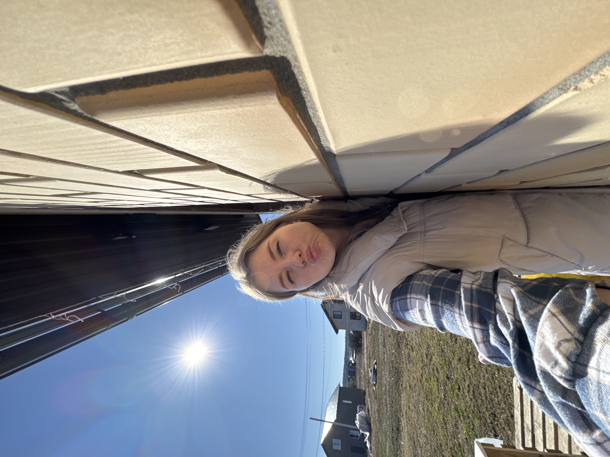 A selfie shows a person between two structures. The person has light-colored hair, wearing a light-colored puffy jacket and a blue and white plaid shirt. The structure above appears to be light brown brick. The structure to the left appears to be dark. In the background, there is a clear blue sky with the sun shining brightly. The location includes houses in a grassy area in Gornyi (Dvurechenskoe S/P), Russia.