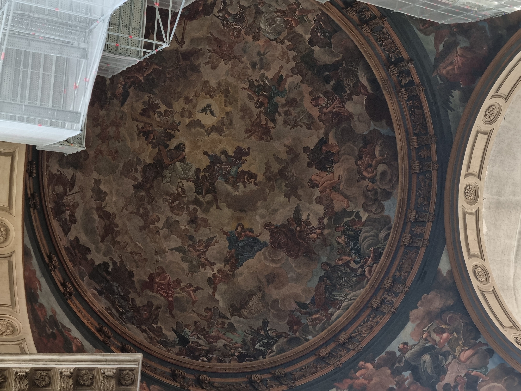 A detailed overhead shot captures the interior of a cathedral, showcasing a vast, circular ceiling fresco surrounded by ornate concentric borders. The fresco depicts a multitude of figures, seemingly angels and other divine beings, amidst swirling clouds in a predominantly sepia palette with hints of blue and red. Portions of the ceiling border are adorned with decorative molding and classical architectural elements. Scaffolding, shrouded in white netting, is visible in the upper left corner, suggesting ongoing restoration or maintenance work. The image composition is slightly off-center, adding to the perspective of looking up at the ceiling from below.