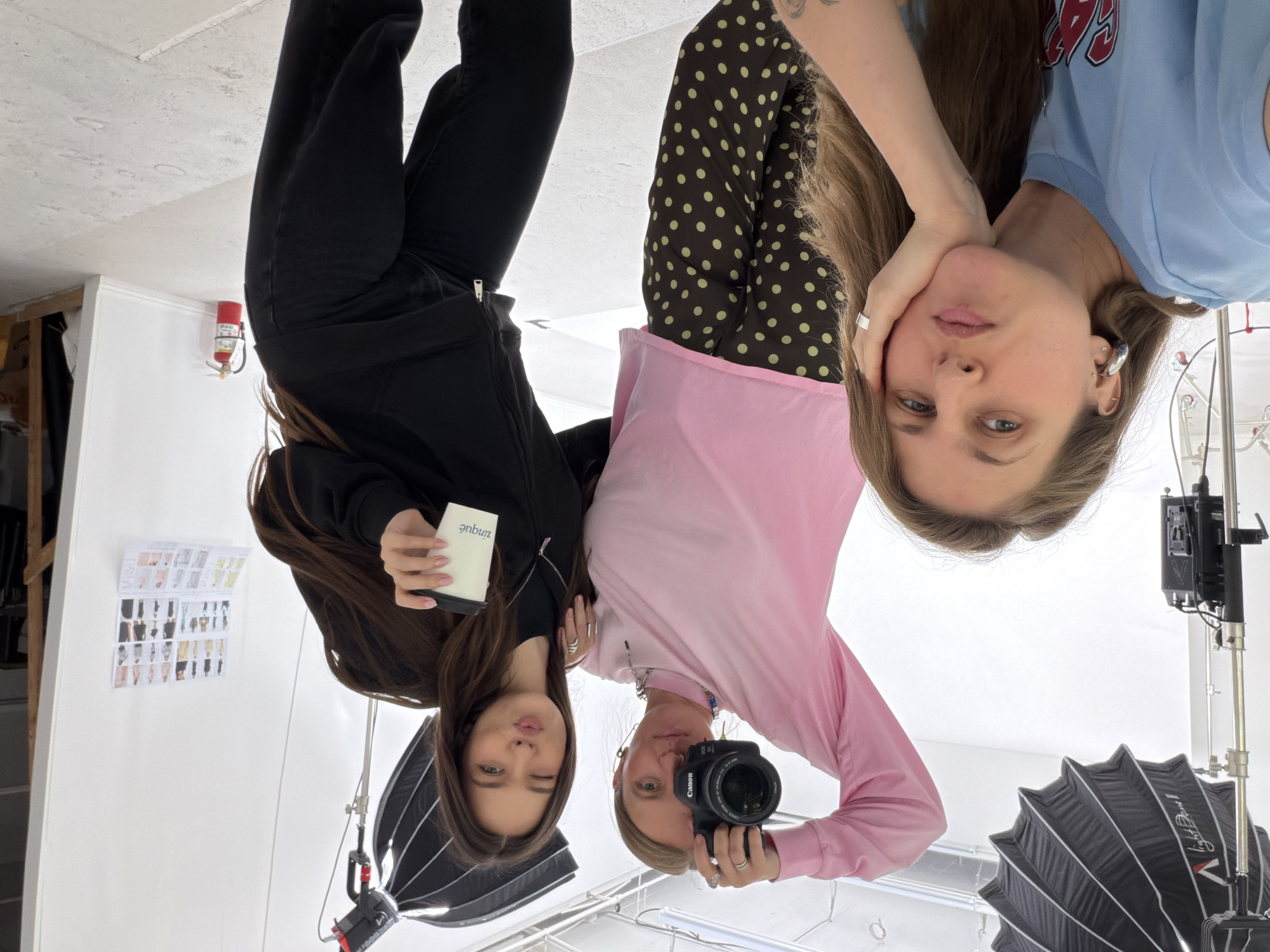 The image captures three individuals, possibly women, within what appears to be a photography studio, likely in Los Angeles, given the location context. The studio environment is suggested by lighting equipment, backdrops, and a mood board displaying images, affixed to a white wall.
The leftmost person wears dark clothing and holds a cup. The individual in the center has a camera and wears a pink top with a pink and brown polka dot pattern underneath. To the right, the third person wears a light blue shirt. They appear to be posing or taking a selfie, with the camera being held by the central figure.
