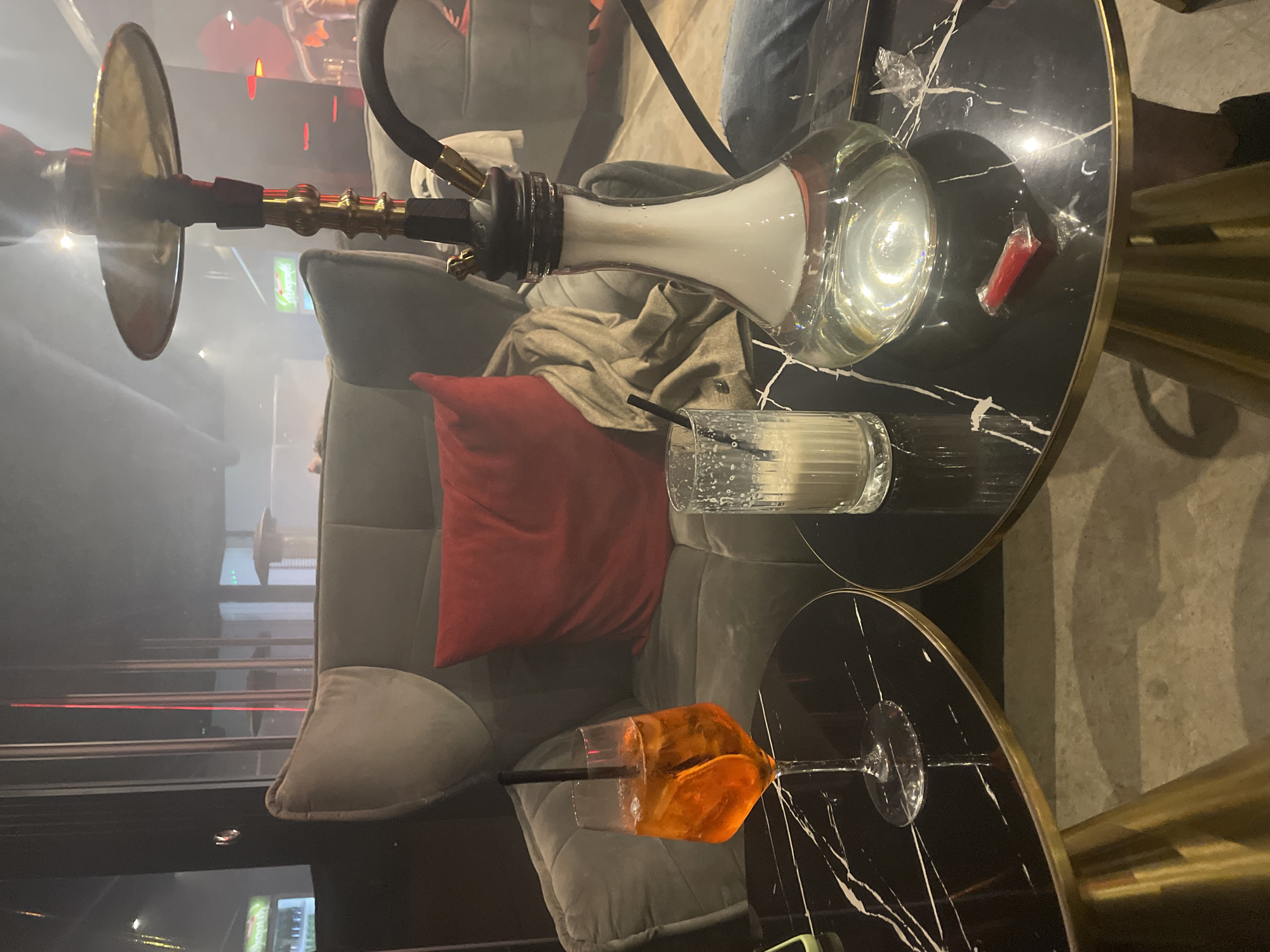 The image shows a lounge setting in Varna, Bulgaria. A lit hookah pipe is positioned on a grey velvet chair with a red pillow. Two round black marble tables with gold trim are present. One table holds a partially consumed white cocktail with ice and a black straw, along with a red wrapper. The other table has a brightly colored orange cocktail with a black straw. The room is dimly lit, and smoke from the hookah is visible in the air.