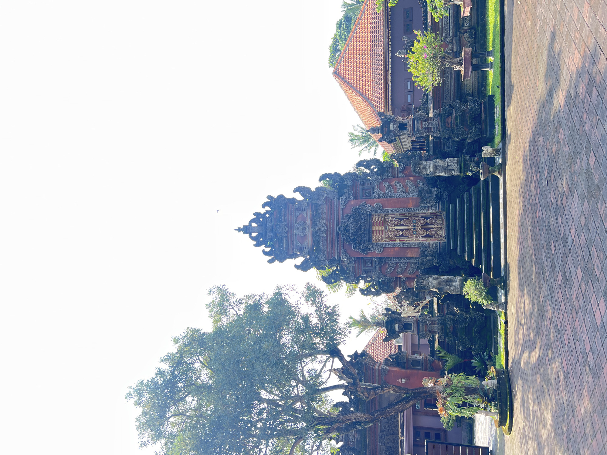 This image depicts a Balinese Hindu temple gate in Sukawati, Indonesia. The gate is constructed of dark stone and ornately carved. It has a series of steps leading up to the gate. To the left of the gate is a large tree with green leaves. To the right of the gate is a building with a red tile roof. A brick walkway extends from the bottom of the image. The sky is pale white.
