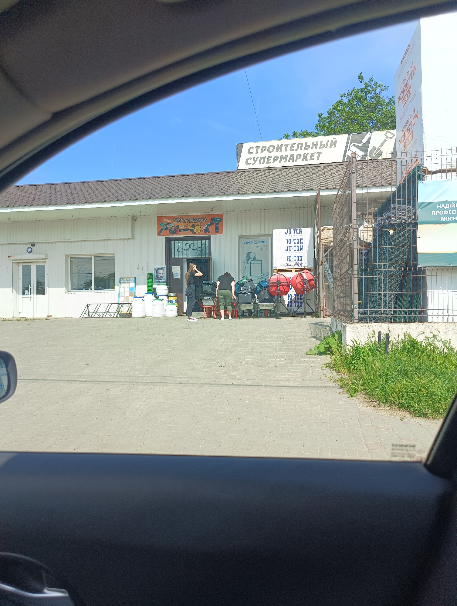 The image, captured from inside a vehicle, presents an exterior view of a building identified as a "Building Supermarket" (СТРОИТЕЛЬНЫЙ СУПЕРМАРКЕТ) in Maiaky, Ukraine, under a clear blue sky. The single-story structure is white with a brown tiled roof.
Near the central entrance, a rectangular sign displays "Stummaxe" alongside images of power tools. To the left of this entrance is a large window and a white double door. Directly in front of the entrance, two individuals are visible. One person, clad in dark clothing, stands facing the entrance, with both hands near their head. The second individual, wearing a dark top and olive green pants, is bent over near a red wheelbarrow on the right side of the entrance.
The paved foreground, constructed of light grey pavers, features various items displayed for sale or storage. These include multiple white plastic barrels and crates, several metal racks, and stacks of building materials labeled "JU-TON." Two red cement mixers on wheels are positioned to the right of the entrance, near a second wheelbarrow. Further right, behind a tall metal mesh fence, are rolls of dark material, likely fencing, and other building supplies. Additional signage on the right includes "ГАЗОБЕТОН-UE PORISTON" and "НАДІЙНИЙ ПРОФЕСІЙНИЙ ЯКІСНИЙ" (Reliable Professional Quality). A sign to the left also mentions "СТРОЙ ЦЕНТР" (Building Center) with "ЛУЧШИЕ ЦЕНЫ!" (Best Prices!).
Green grass and weeds grow along the right edge of the paved area, adjacent to the metal fence. The background reveals a cluster of green trees against the blue sky. The left side of the image shows a portion of the vehicle's interior, including a dark dashboard and a side mirror.