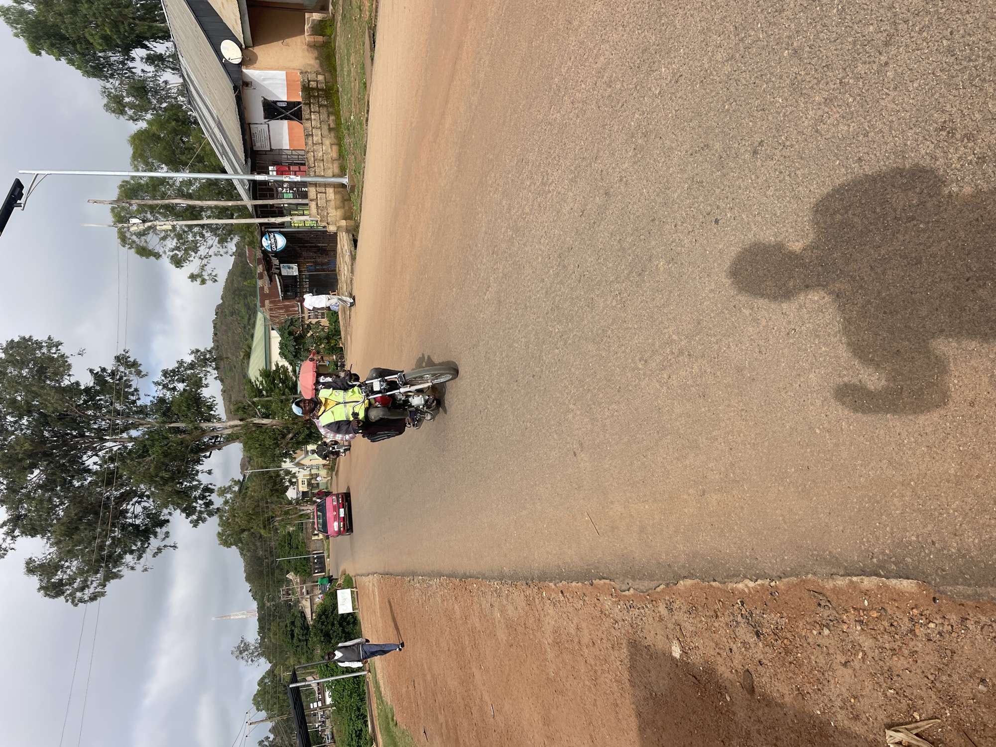 In Gura Top, Nigeria, on a paved road with a dirt shoulder, a man on a motorcycle wearing a reflective vest rides forward, casting a shadow on the asphalt. A red car is also visible further down the road, as is a man in a white shirt walking on the dirt shoulder. In the background, buildings and trees line the street, under a cloudy sky. A solar-powered street light stands tall on the side of the road.