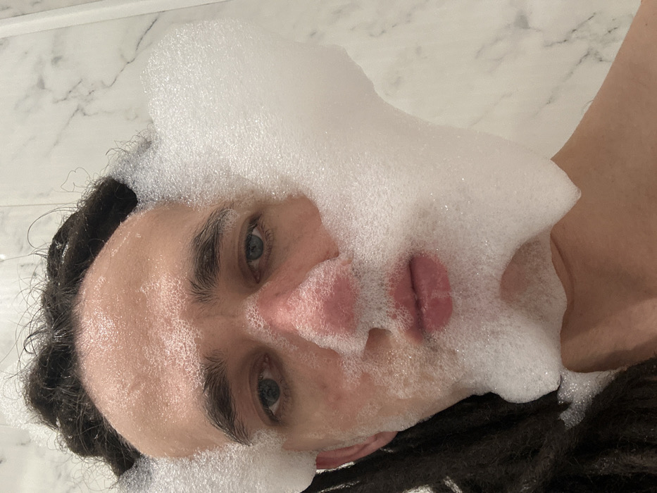 A person lies on their side with their face covered in soapy foam. The foam obscures much of their face, except for the eyes, nose, and lips. Their dark curly hair is visible around the edges of their face. The background appears to be a tiled surface, likely a bathroom. The image is tilted, giving an unusual perspective. The overall scene suggests a playful or relaxed moment during a bath or shower.