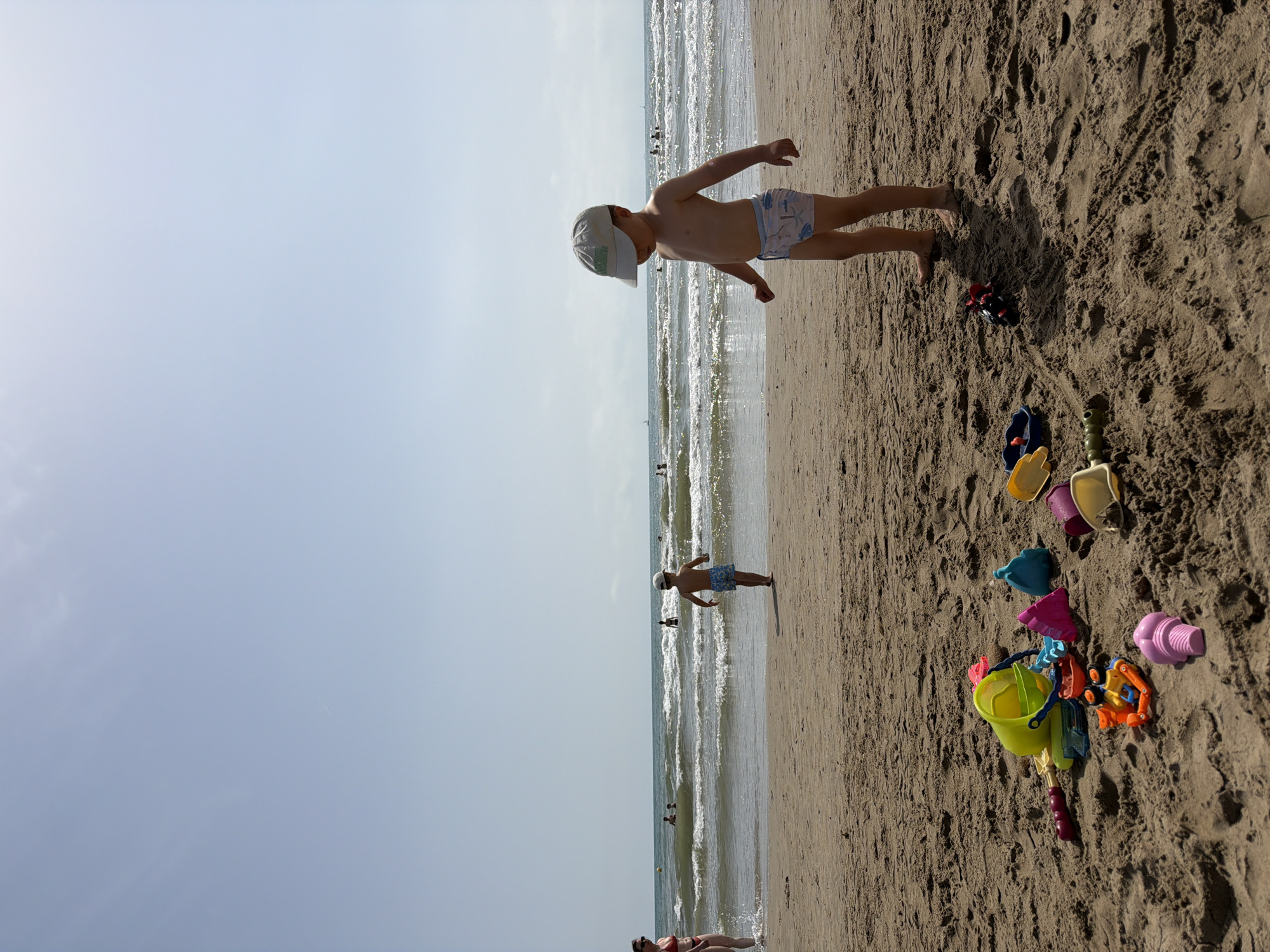 In an outdoor scene on a sandy beach, under a clear sky, a group of children and adults are present. In the foreground, a collection of brightly colored plastic beach toys, including buckets, shovels, and sand molds, are scattered on the sand. Two young boys are visible. One boy, wearing blue patterned swim trunks and a light-colored cap, stands facing away from the camera, looking towards the ocean. He is barefoot. Another boy, further in the background, is running towards the ocean. A woman in a red bikini stands near the water's edge, facing left. The ocean, with moderate waves, stretches out to the horizon, where several people can be seen swimming and playing in the water. The sand is a light brown color, with a textured surface. The overall atmosphere suggests a summer day at the beach.