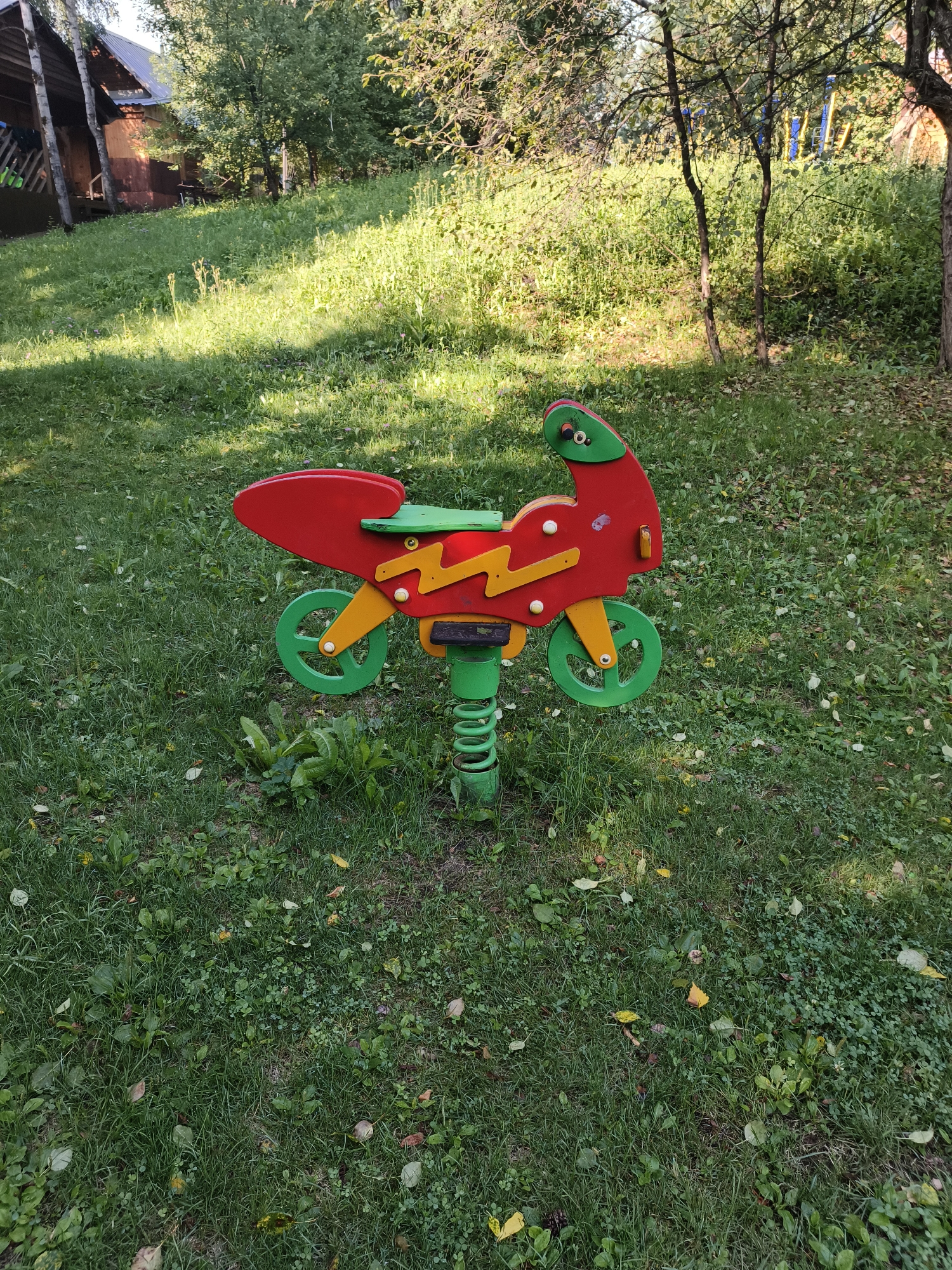 A red and green spring-mounted toy motorcycle is positioned on a grassy lawn in a wooded area. The motorcycle has yellow zigzag decorations on its sides and green wheels. In the background, a rustic wooden building with a gray roof is visible among trees. The ground is covered in green grass with some scattered fallen leaves. The lighting suggests it is daytime.