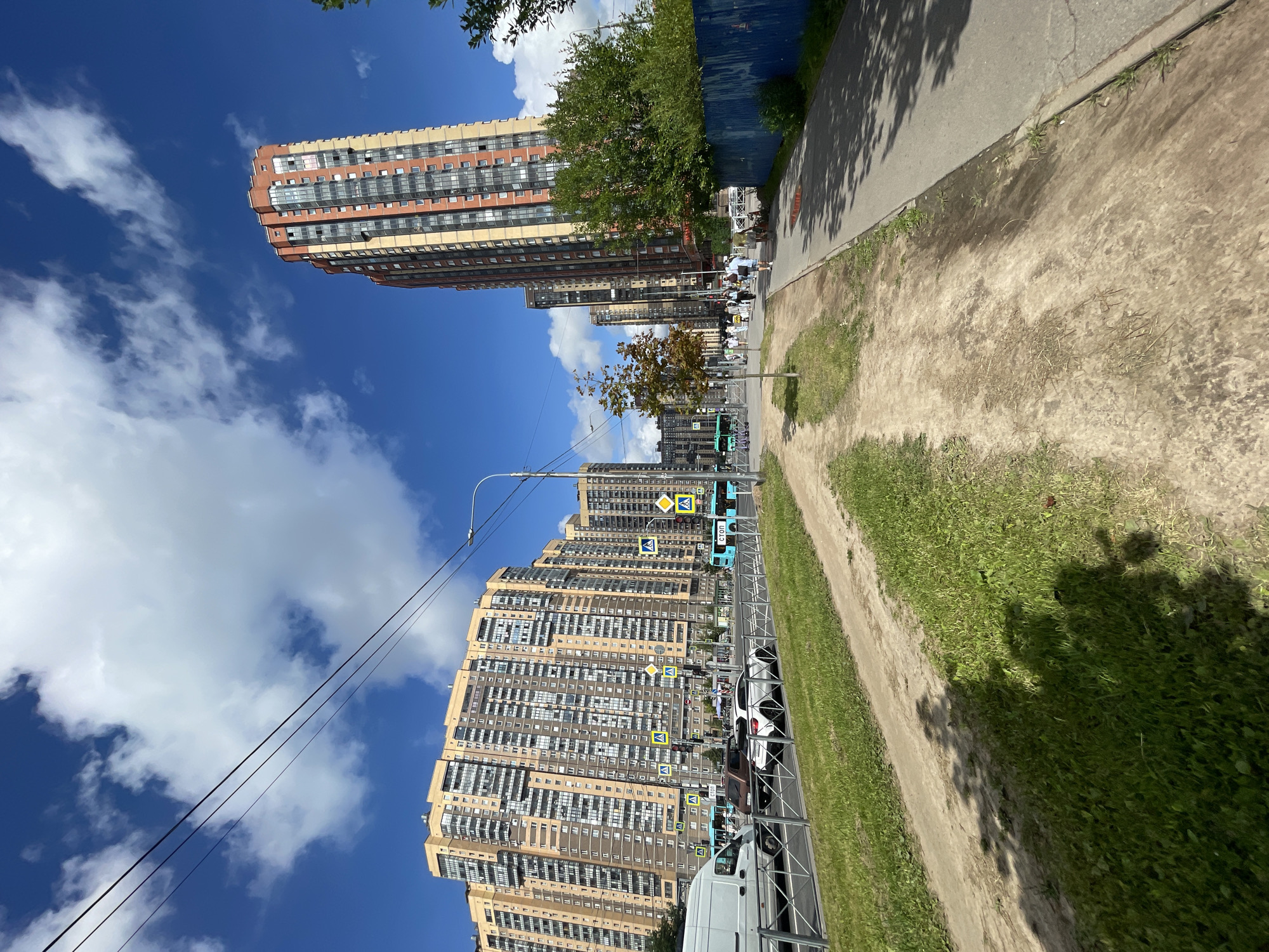 The image depicts an urban scene in an unknown location in Russia. The foreground features a dirt path running alongside a paved sidewalk, bordered by a grassy area. In the background, a multi-lane road is visible with various vehicles, including cars, a white van, and blue buses. Several pedestrians are walking along the sidewalk and crossing the street. Dominating the skyline are tall, modern apartment buildings with light-colored facades and numerous windows. The sky is blue with scattered white clouds, and power lines are strung across the upper portion of the image. A tall, orange and yellow building stands out among the residential blocks.