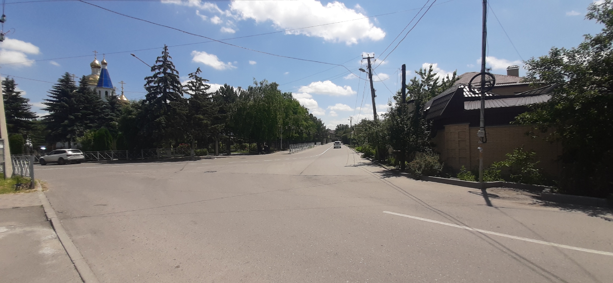 The image captures a sunny day in Yessentukskaya, Russia, with a wide asphalt road dominating the foreground. On the left side of the road, a white SUV is parked near a metal fence. Behind the SUV, a white building with golden domes and a blue spire, likely a church, is visible amidst a cluster of tall evergreen trees. The right side of the road features a brick fence and a dark-tiled roof of a building, partially obscured by lush green trees and bushes. The sky is a clear blue, dotted with fluffy white clouds. Power lines are strung across the scene, supported by wooden poles. Further down the road, a few more cars are visible in the distance, and the road appears to lead into a more densely vegetated area. The overall environment suggests a suburban or semi-rural setting with a mix of residential buildings and natural greenery.