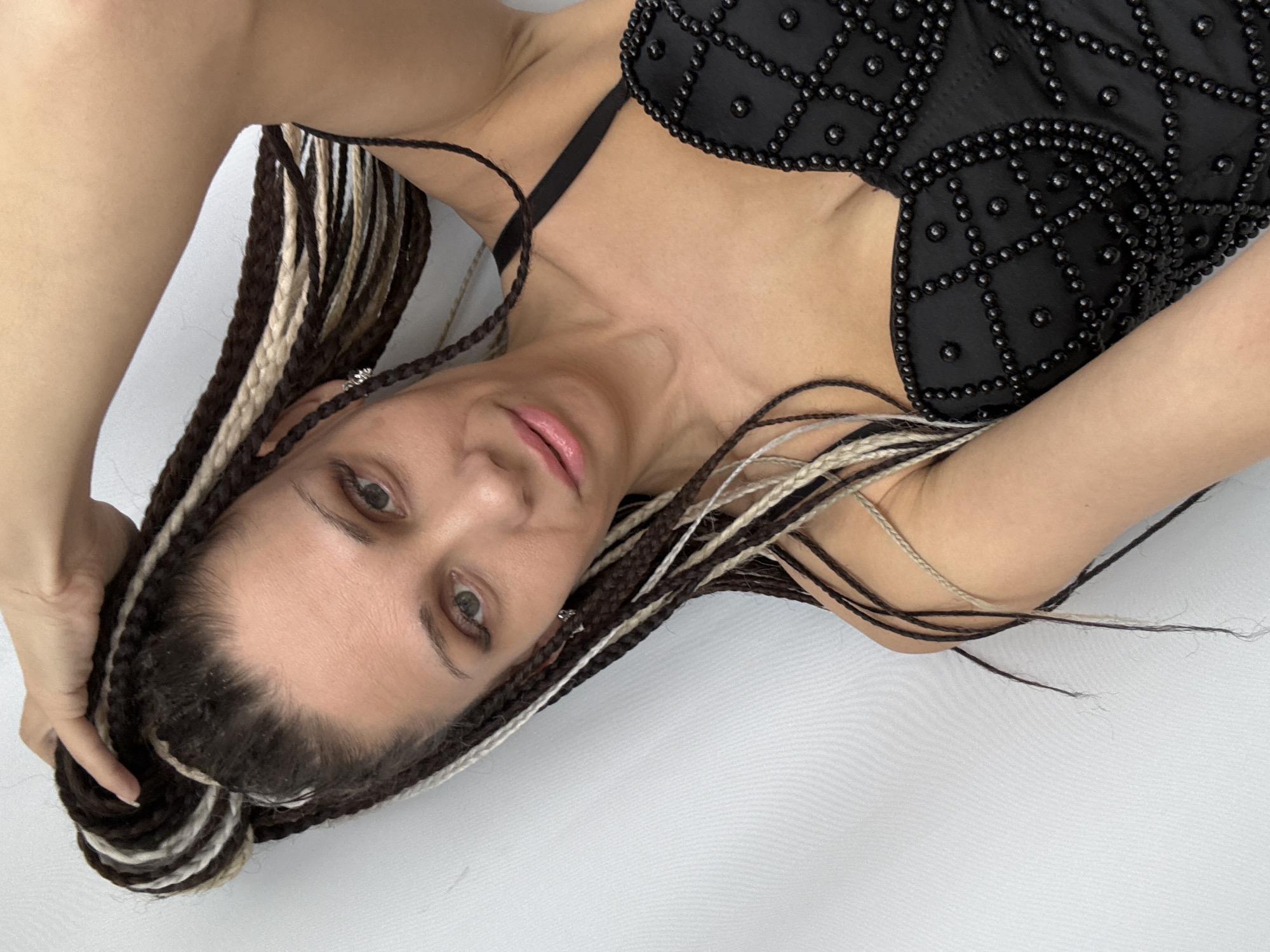 A woman with dark hair braided with blonde extensions is laying down on a white surface, possibly fabric. She is wearing a black top with decorative beading. The photo appears to be taken from a high angle, looking down at the subject. The background is plain white. There is no indication that the image was taken in Rusanovo, Russia.