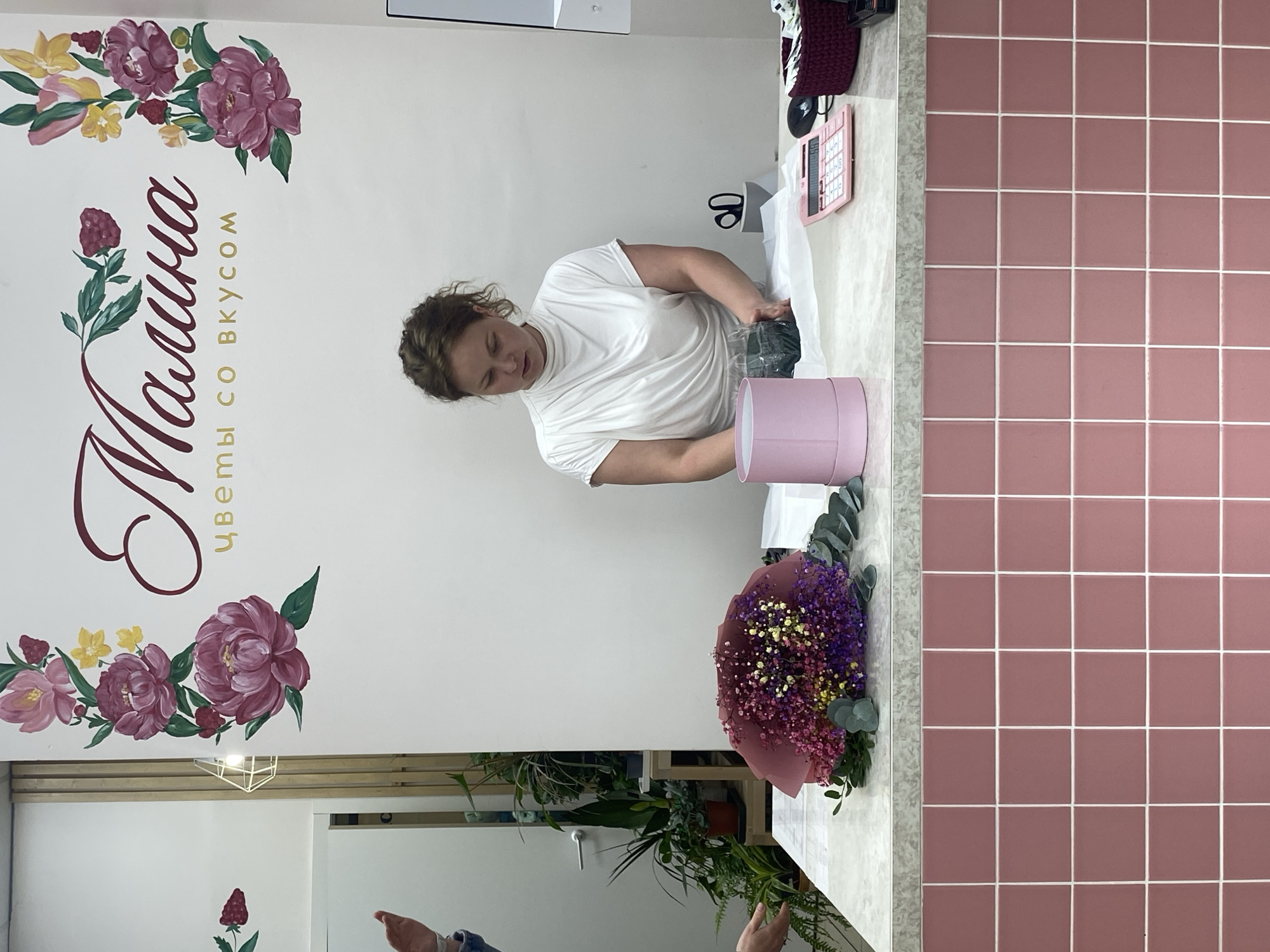 The photograph depicts a woman in a white t-shirt working at a floral shop, as indicated by the floral arrangements, supplies, and business signage present in the scene. She appears to be arranging or packing a bouquet of flowers into a pink cylindrical box, with wrapping paper and other floral arrangement materials surrounding her.
The background features a wall adorned with a floral design and the business name "Малина" in Russian, along with the phrase "цветы со вкусом" (flowers with taste). There are plants in the background.
The shop has a countertop with a calculator, and the countertop runs adjacent to a wall covered in pink tiles.
The location is indicated by the Russian signage, which, when translated, suggests this business could be located in a Russian-speaking area, possibly Yekaterinburg, Russia.