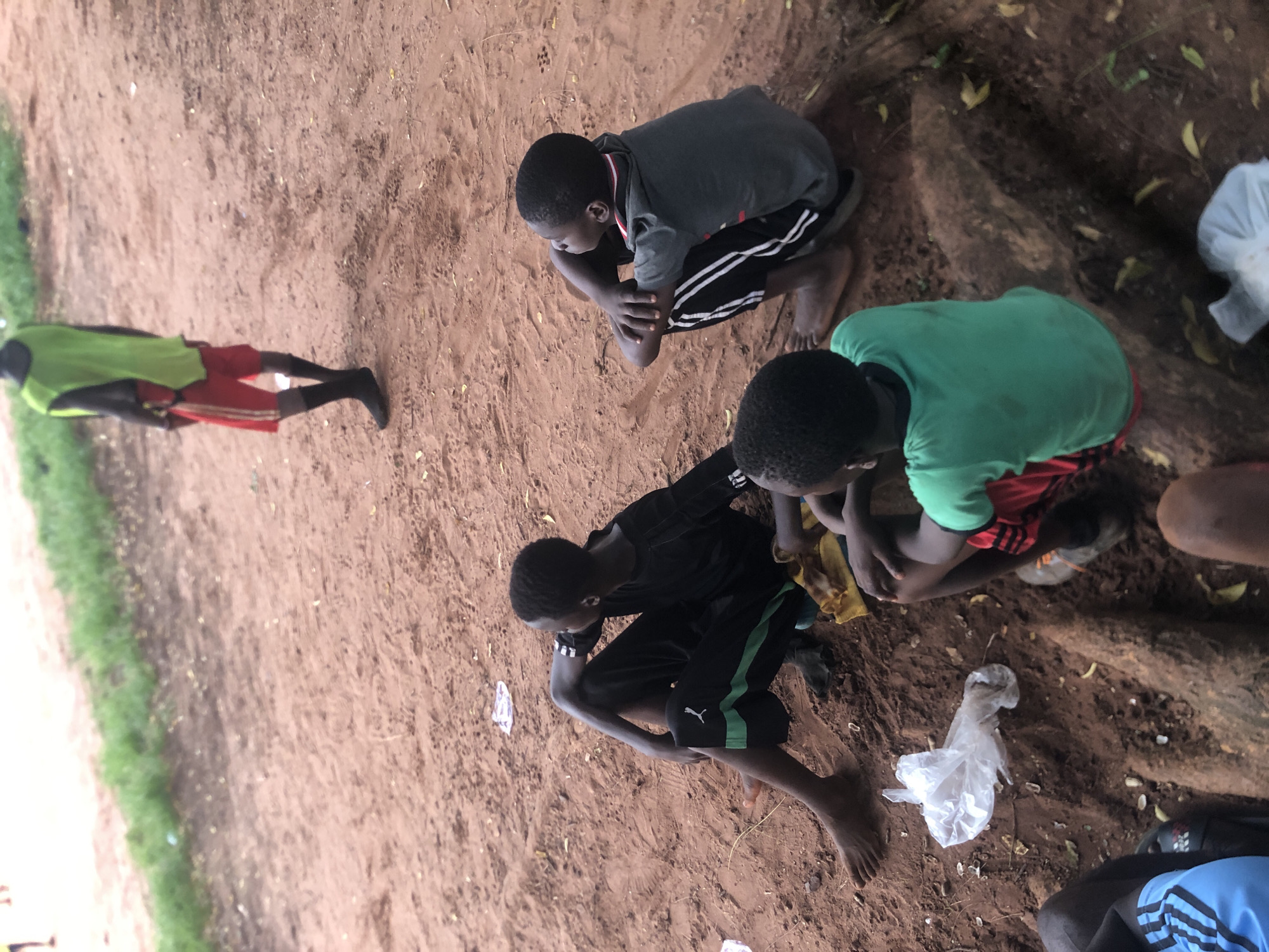 The image shows a group of boys in Ukwu-Abwa, Nigeria, on reddish-brown soil near a grassy area. Three boys are crouched close together in a circle, while another boy stands further away. The boys are dressed in athletic clothing, including t-shirts, shorts with stripes, and one boy is wearing leggings. There are plastic bags and some leaves scattered on the ground. They are gathered near a tree root or log. The area appears to be outdoors with dirt underfoot.