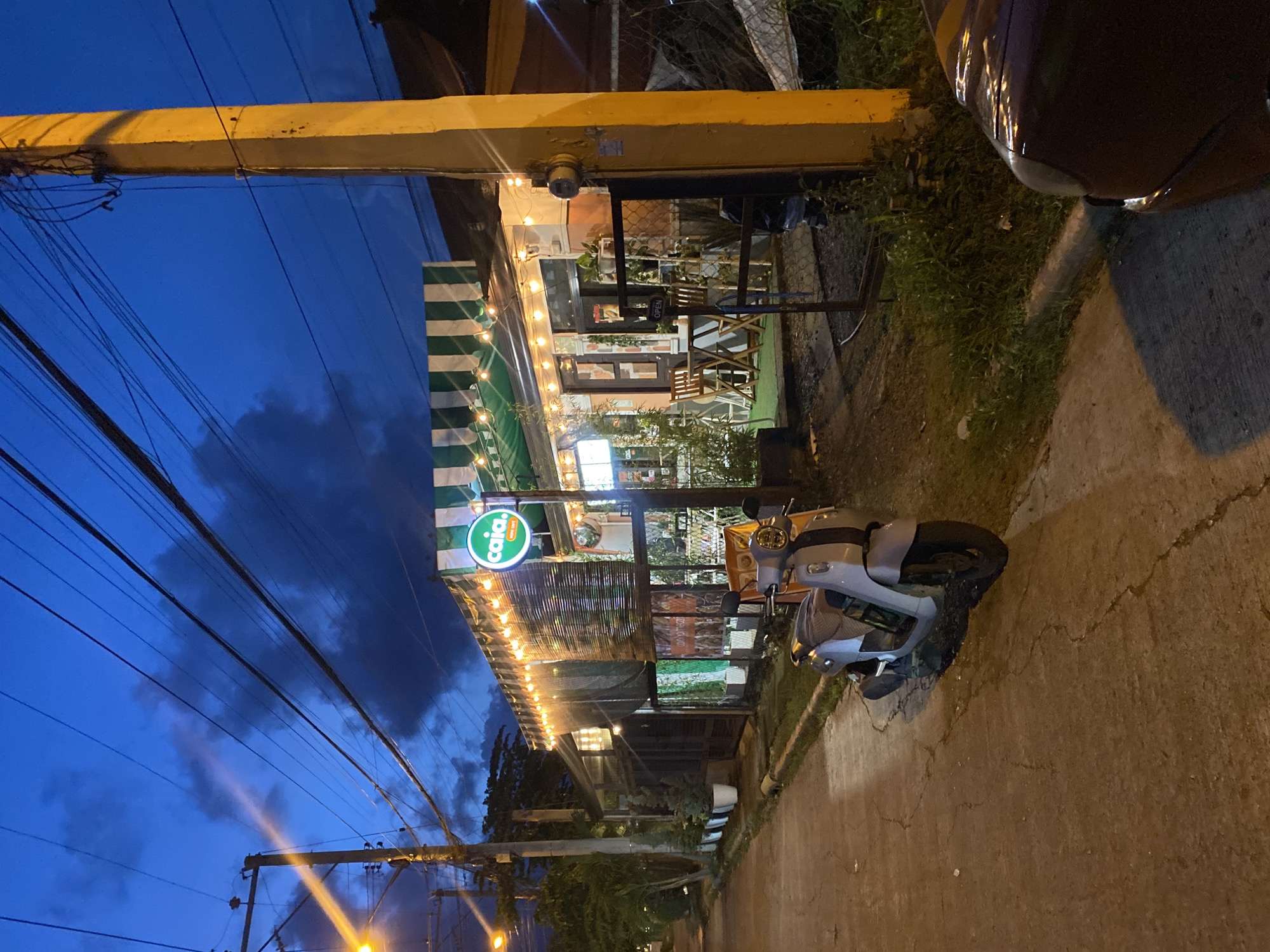 The image depicts a street scene at dusk in Davao City, Philippines. On the right side of the frame, a yellow utility pole stands prominently, casting a shadow. A dark-colored vehicle's fender is visible in the lower right corner. A light-colored scooter is parked on the concrete pathway in the foreground.
Dominating the center of the image is a building with a green and white striped awning, adorned with string lights. A circular sign with the word "caia." is affixed to the awning. The building appears to be a cafe or restaurant, with tables and chairs visible through a fence and glass doors. A "OPEN" sign is displayed on the gate. Behind the building, dense foliage and trees can be seen. Electrical wires crisscross the deep blue sky, where scattered clouds are present. The overall atmosphere suggests a quiet evening in a developing urban area.