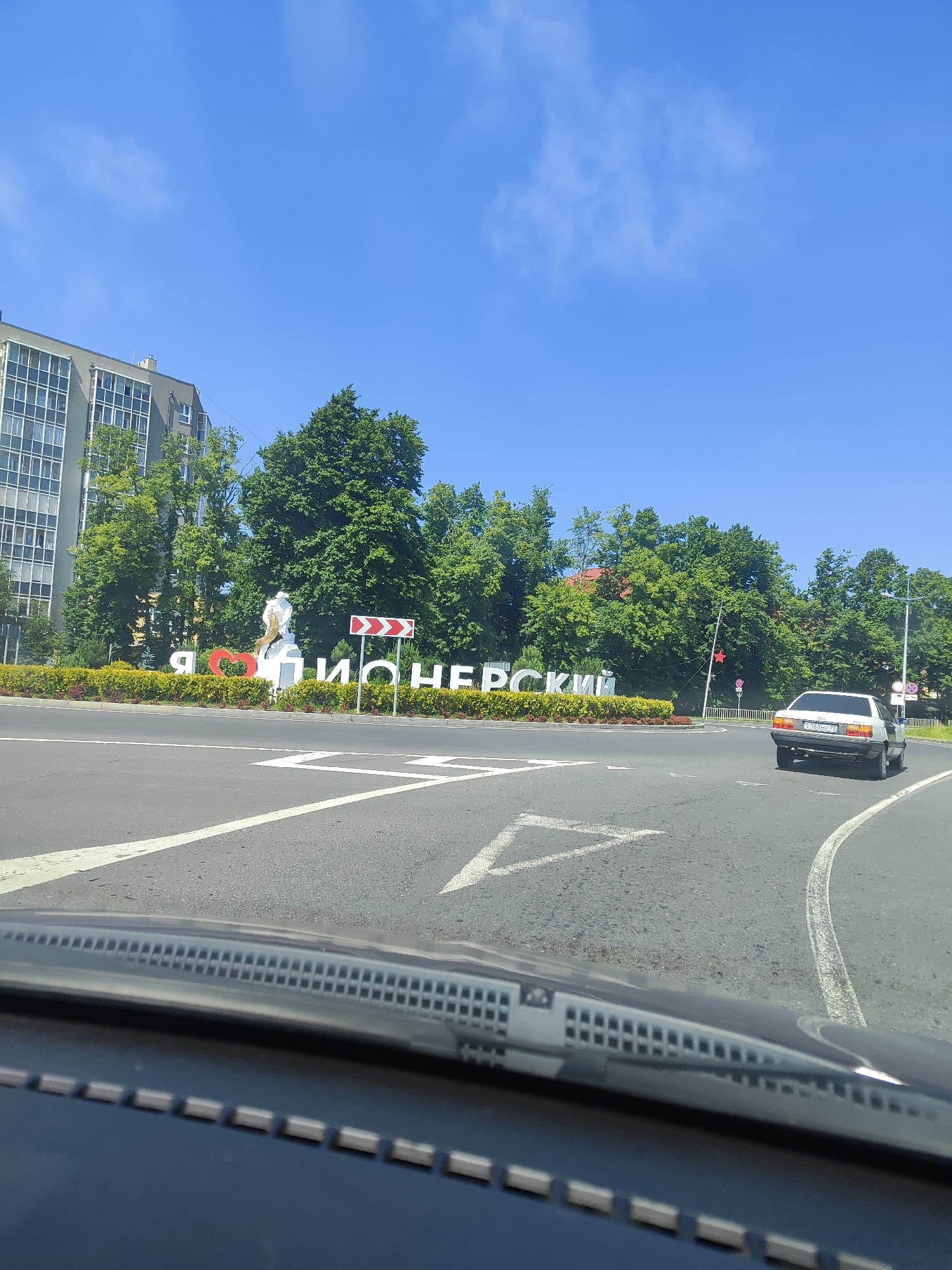 The image depicts a street scene in Pionersky, Russia, on a clear, sunny day. In the foreground, the dashboard and windshield of a vehicle are visible, suggesting the photo was taken from a car. The road ahead curves to the right, with white lane markings and an arrow indicating direction.
A prominent feature in the middle ground is a large sign with white letters spelling out "Я ЛЮБЛЮ ПИОНЕРСКИЙ" (I Love Pionersky), with a red heart in place of the word "люблю" (love). Behind the sign, there is a sculpture or monument and a well-maintained hedge.
To the left of the sign, a multi-story building with many windows is visible, partially obscured by lush green trees. The trees form a dense canopy across the background.
On the right side of the road, a white car, possibly a sedan, is driving away from the camera. Further in the background, more trees and some distant structures are visible. Traffic signs and streetlights can also be observed. The sky is a bright blue with a few wispy clouds.